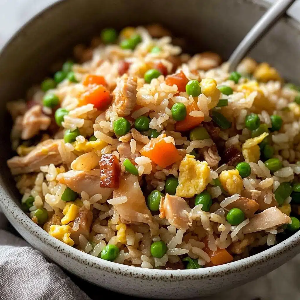 Chicken Fried Rice, quick fried rice recipe, easy weeknight dinner, flavorful fried rice, homemade fried rice A close-up view of a bowl filled with fried rice that has several layers and colors: at the base, white and light brown rice grains mixed with small cubes of orange carrots and green peas; scattered on top are pieces of light brown cooked chicken, tiny yellow scrambled egg bits, and small dark red-brown meat cubes; finishing with bright green chopped spring onions sprinkled over the rice. The bowl is light gray with subtle dark specks, and a silver spoon is partially visible, standing upright in the bowl. The surface under the bowl is a white marbled texture with a dark gray cloth.