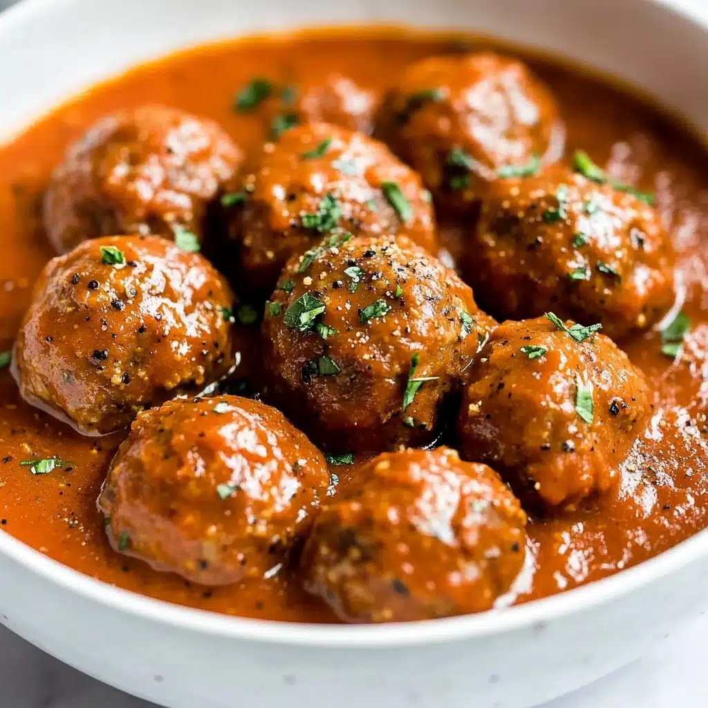 Saucy Beef and Lentil Meatballs, hearty meatball recipe with lentils, easy wholesome dinner ideas, flavorful beef meatballs with coconut milk, healthy comfort food recipes The image shows round meatballs covered in a thick tomato sauce inside a white bowl. The sauce is smooth and reddish-orange in color, coating the meatballs evenly. The meatballs have a brown color with a slightly bumpy texture, and small green herb pieces are sprinkled on top, adding a fresh touch. Black pepper specks are visible on the meatballs and sauce, giving them extra texture. The bowl sits on a white marbled surface.