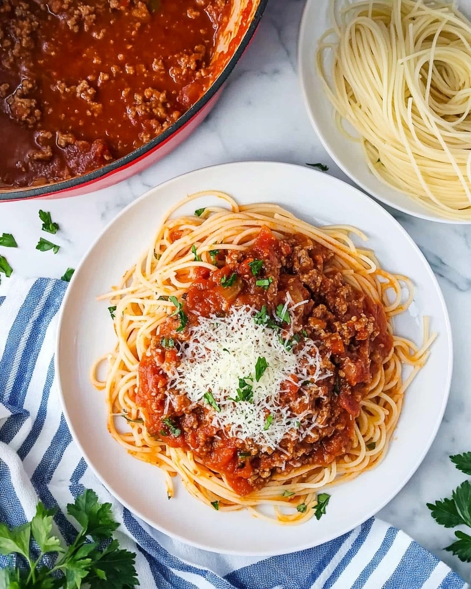 Turkey Spaghetti with Tomato Sauce and Vegetables, healthy turkey pasta, quick weeknight dinner, veggie-packed spaghetti, light comfort food A white plate holds a dish with three main layers: a bottom bed of light brown cooked spaghetti noodles, a thick and chunky bright red tomato meat sauce covering the noodles on top, and a sprinkling of grated pale yellow cheese along with chopped green herbs scattered over the sauce. The plate sits on a wooden surface with some green herb leaves around, and a striped cloth with dark blue and white colors is partly visible in the background on one side. A shiny silver pot with more spaghetti inside is placed in the background on the right side.