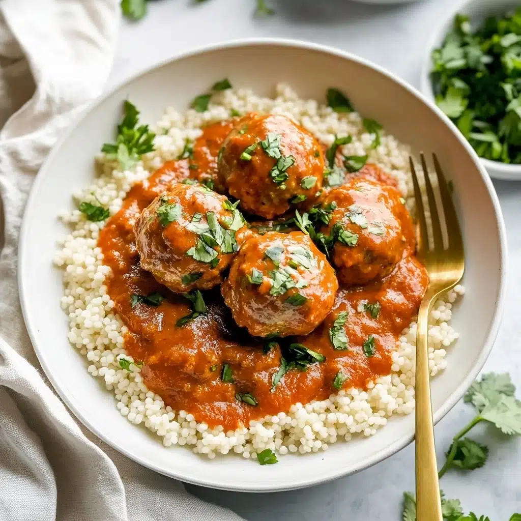 Saucy Beef and Lentil Meatballs, hearty meatball recipe with lentils, easy wholesome dinner ideas, flavorful beef meatballs with coconut milk, healthy comfort food recipes A white bowl holds a bed of small, pearl-like couscous grains that are off-white and slightly shiny. On top, there are five medium-sized meatballs covered in a smooth, thick orange-red sauce. The sauce looks creamy and has small green herb pieces sprinkled over it. Some fresh green herbs also decorate around the meatballs on the couscous. A gold fork is partially visible, placed inside the bowl on the right side. The bowl sits on a white marbled surface with a white cloth nearby and some green herbs in a small white bowl in the background.