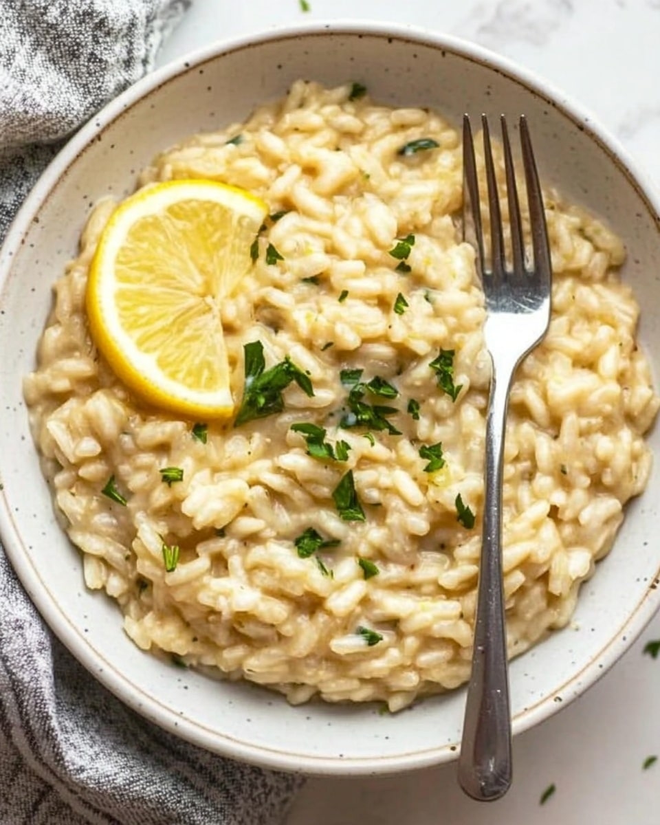 One Pot Lemon Orzo Pasta, Lemon Orzo Pasta, Easy Lemon Orzo Recipe, Creamy Lemon Pasta, One Pot Pasta Dinner A white bowl filled with creamy risotto, creamy light yellow in color with small green herb pieces mixed throughout. On the left side inside the bowl, two thin lemon slices rest against the risotto. A silver fork is placed inside the bowl on the right side, partially submerged in the risotto. The bowl sits on a white marbled surface with a grey and white cloth napkin partially visible at the bottom left corner.