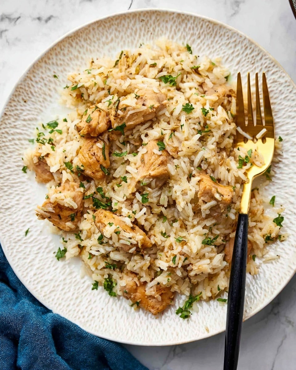 The image shows a white plate with a textured rim filled with a mixture of cooked rice and pieces of brownish-yellow chicken. The chicken pieces are scattered unevenly throughout the rice, which is soft and slightly clumped. Small green flecks of chopped herbs are sprinkled on top, adding a touch of color. A gold fork with a black handle rests on the right side of the plate, partly touching the food. The plate sits on a white marbled surface with a blue cloth partially visible on the left side of the image. photo taken with an iphone --ar 4:5 --v 7