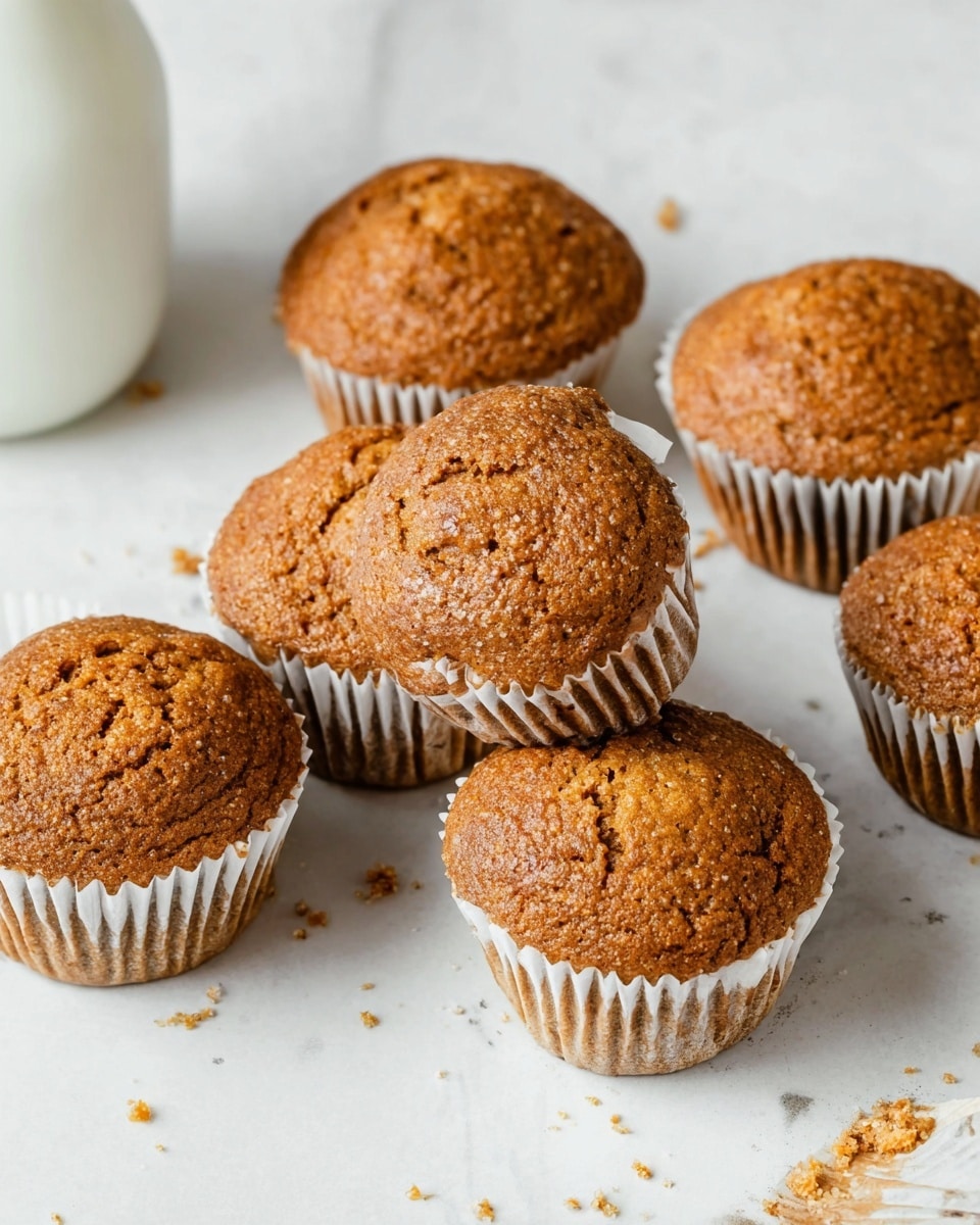 The image shows six brown muffins with slightly cracked tops and a rough texture, placed on a white marbled surface. Each muffin is wrapped in a white paper cup, and there are some small crumbs scattered around them. The muffins have a warm, golden-brown color, and one muffin is tilted on its side, showing its ridged paper cup. A white bottle or jar is partially visible in the top left corner. Photo taken with an iphone --ar 4:5 --v 7