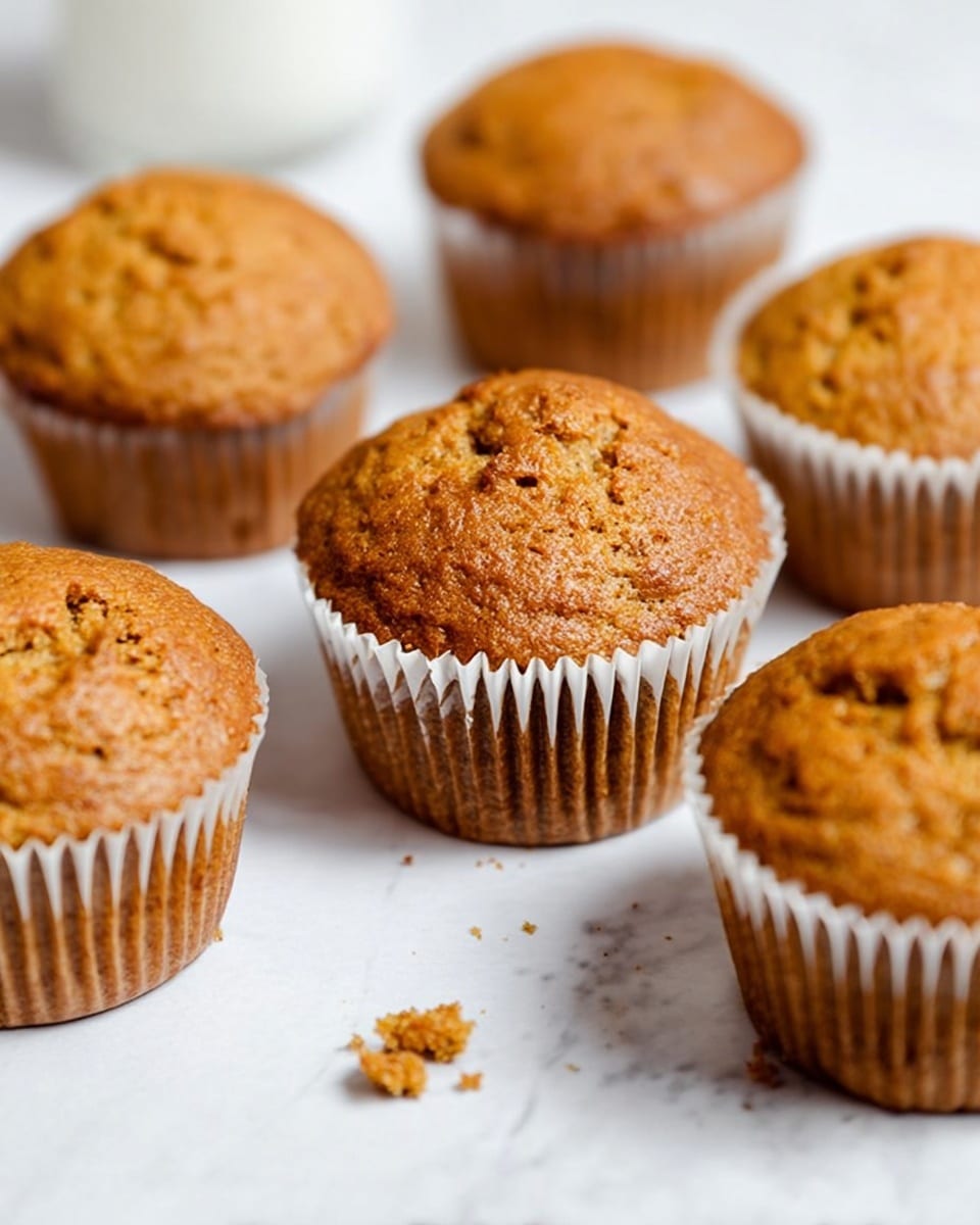 The image shows six golden brown muffins placed on a white marbled surface, with one muffin in the center and others arranged around it. The muffins have a slightly cracked top and a soft, textured surface. Each muffin is wrapped in a white paper liner that contrasts with the warm brown color of the muffins. There are small crumbs scattered near the closest muffin, adding a touch of realism. The background is softly blurred, making the muffins the clear focus of the image. photo taken with an iphone --ar 4:5 --v 7