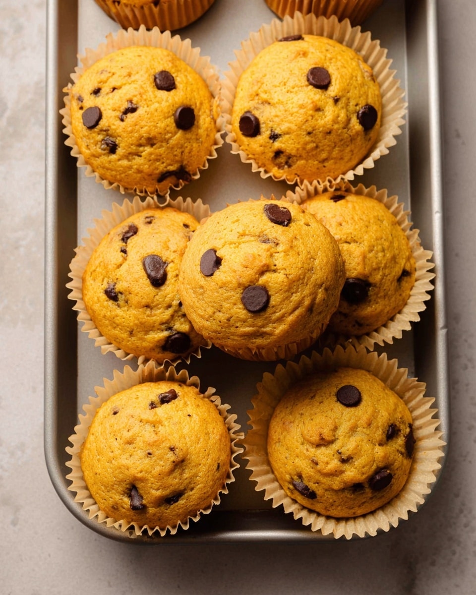 Seven pumpkin chocolate chip muffins with a golden orange color and a slightly cracked top are arranged on a white marbled textured surface inside a silver baking pan. The muffins each sit in a light beige paper liner that holds their shape, and small dark chocolate chips are scattered throughout the muffin tops and visible along the sides. The muffins have a soft and slightly bumpy texture that shows they are baked to a light fluffiness, filling most of the liner space with some slight doming at the top. Photo taken with an iphone --ar 4:5 --v 7