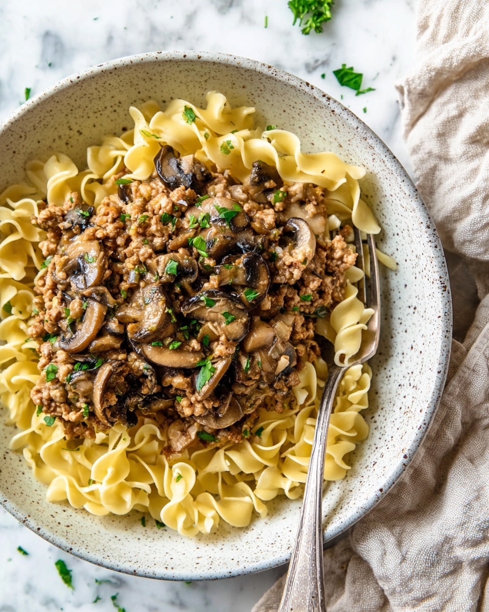 A white speckled bowl holds three main layers of food. The bottom layer is light yellow egg noodles with a soft, curled texture that fill most of the bowl. On top is a thick layer of ground meat sauce mixed with sliced dark brown mushrooms and small pieces of translucent cooked onions, all coated in a light brown creamy sauce. Small green parsley leaves are scattered on the meat mixture, adding a fresh touch. A silver fork rests on the right side of the bowl, partially inside it, and a beige linen cloth is seen nearby on a white marbled surface. Photo taken with an iphone --ar 4:5 --v 7