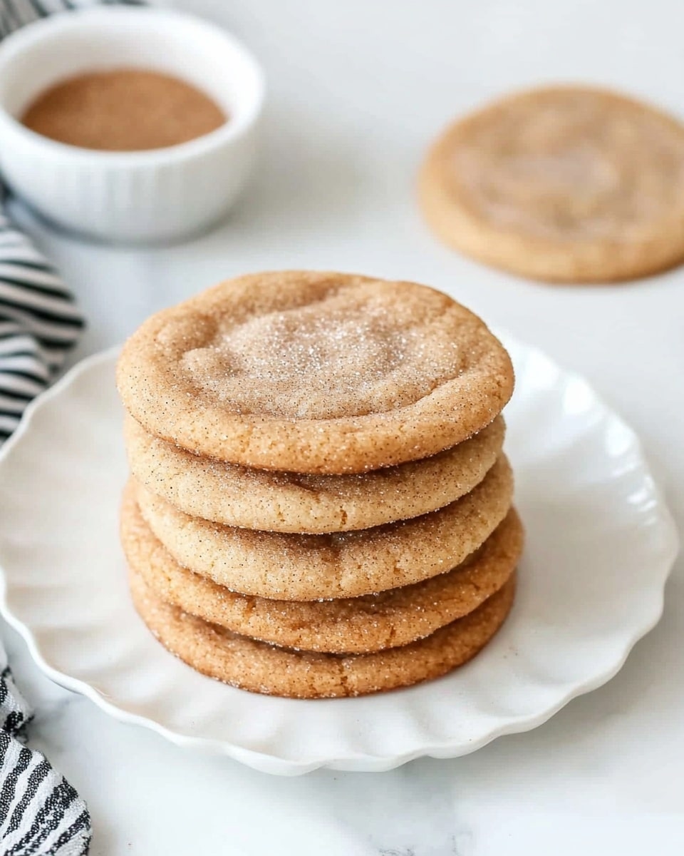 A stack of eight round cinnamon sugar cookies with a light brown color and a fine sugar coating sits in the center of a white scalloped plate, showing a soft texture with some cracks on top. Behind the stack, a single cookie lies flat, slightly darker in some spots. The plate rests on a white marbled surface next to a white bowl filled with cinnamon sugar and a black-and-white striped cloth on the left side. photo taken with an iphone --ar 4:5 --v 6.1