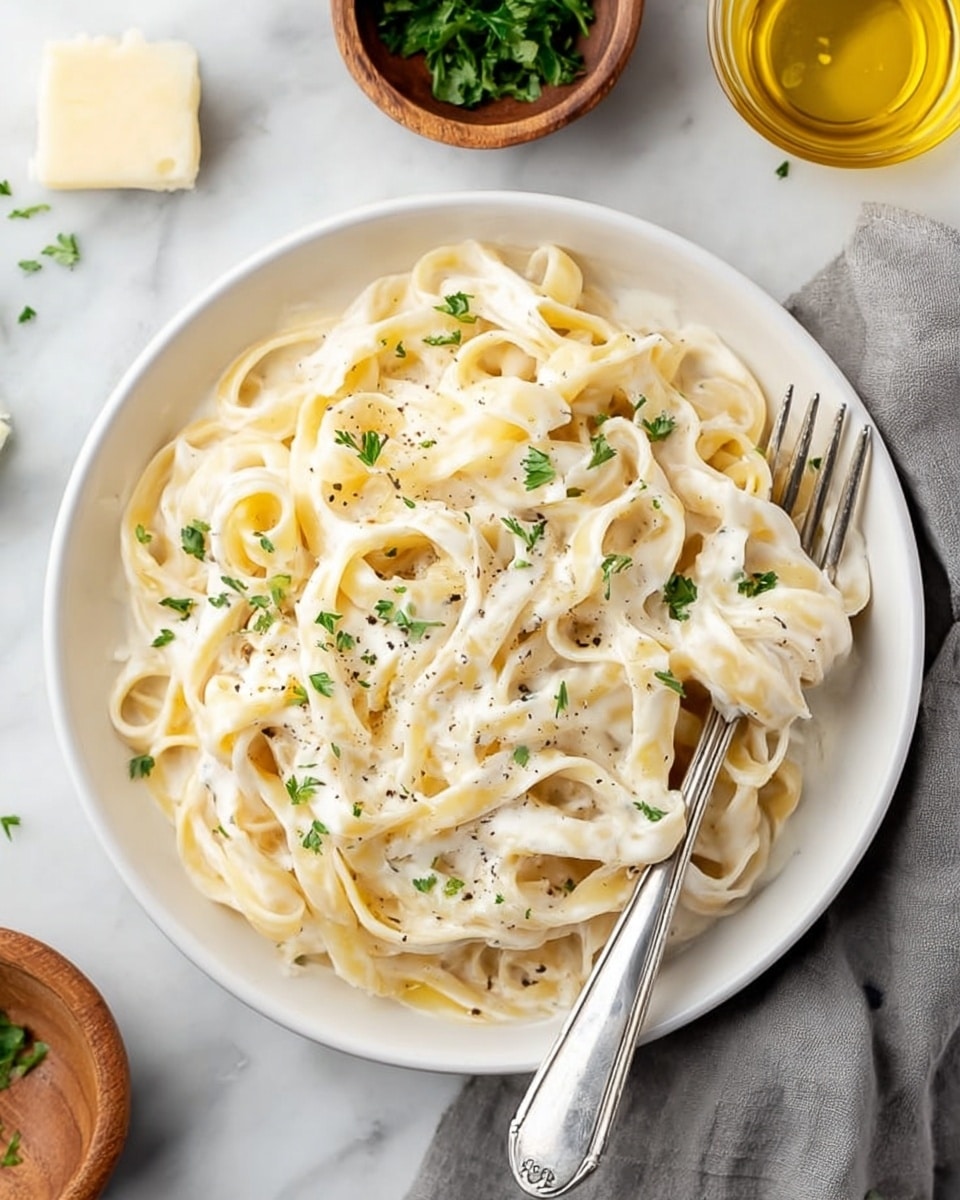 A white bowl filled with creamy fettuccine pasta covered in a smooth white sauce, garnished with small green parsley pieces scattered on top. A silver fork rests on the right side of the bowl, with some pasta gently wrapped around it. The bowl is placed on a white marbled surface, with a grey cloth napkin nearby and a small wooden bowl with green herbs partially visible in the upper left corner. There is also a small chunk of pale yellow butter near the upper left edge and a glass container with golden olive oil at the bottom right corner. The overall colors are soft and light, emphasizing the creamy texture and fresh garnish of the dish photo taken with an iphone --ar 4:5 --v 7