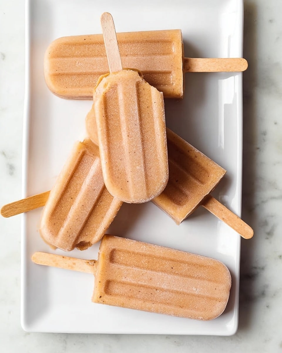 Five light brown popsicles with smooth texture are arranged on a white rectangular plate placed on a white marbled surface. Four popsicles lie flat with wooden sticks pointing sideways, while the center popsicle rests on top, angled slightly, with a bite taken out of its top left corner showing a denser inside. The wooden sticks are light beige and stick out evenly from each popsicle. The photo is bright and clear, showing slight frost on the popsicles. photo taken with an iphone --ar 4:5 --v 7