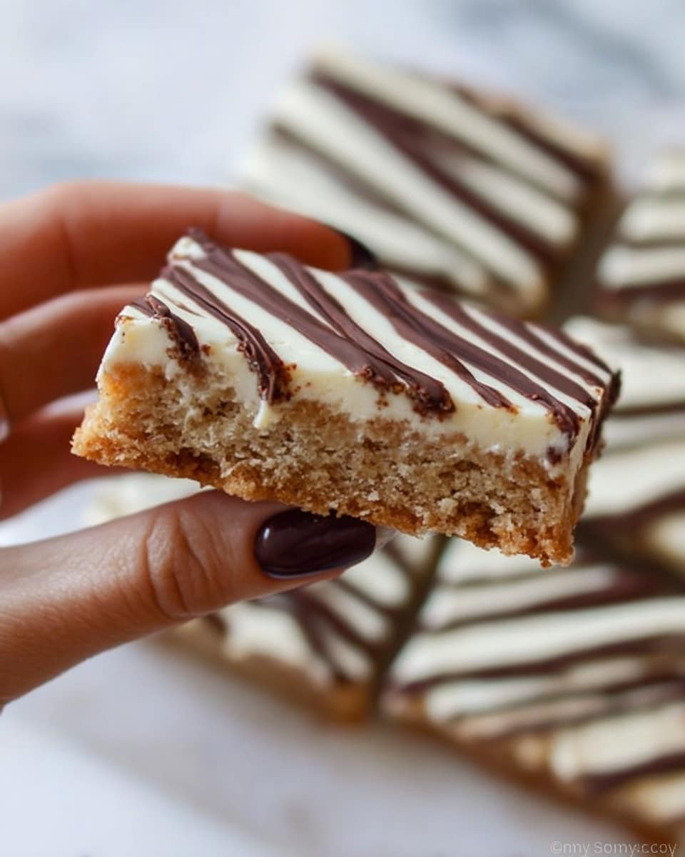 A close-up of a hand holding a rectangular cookie bar with two main layers. The bottom layer is thick and light brown with a crumbly texture and small visible chunks inside. The top layer is smooth and creamy white, evenly spread over the cookie base. Thin dark brown chocolate lines are drizzled across the top layer in parallel, creating a striped pattern. The background shows more cookie bars blurred out on a white marbled surface. Photo taken with an iphone --ar 4:5 --v 7