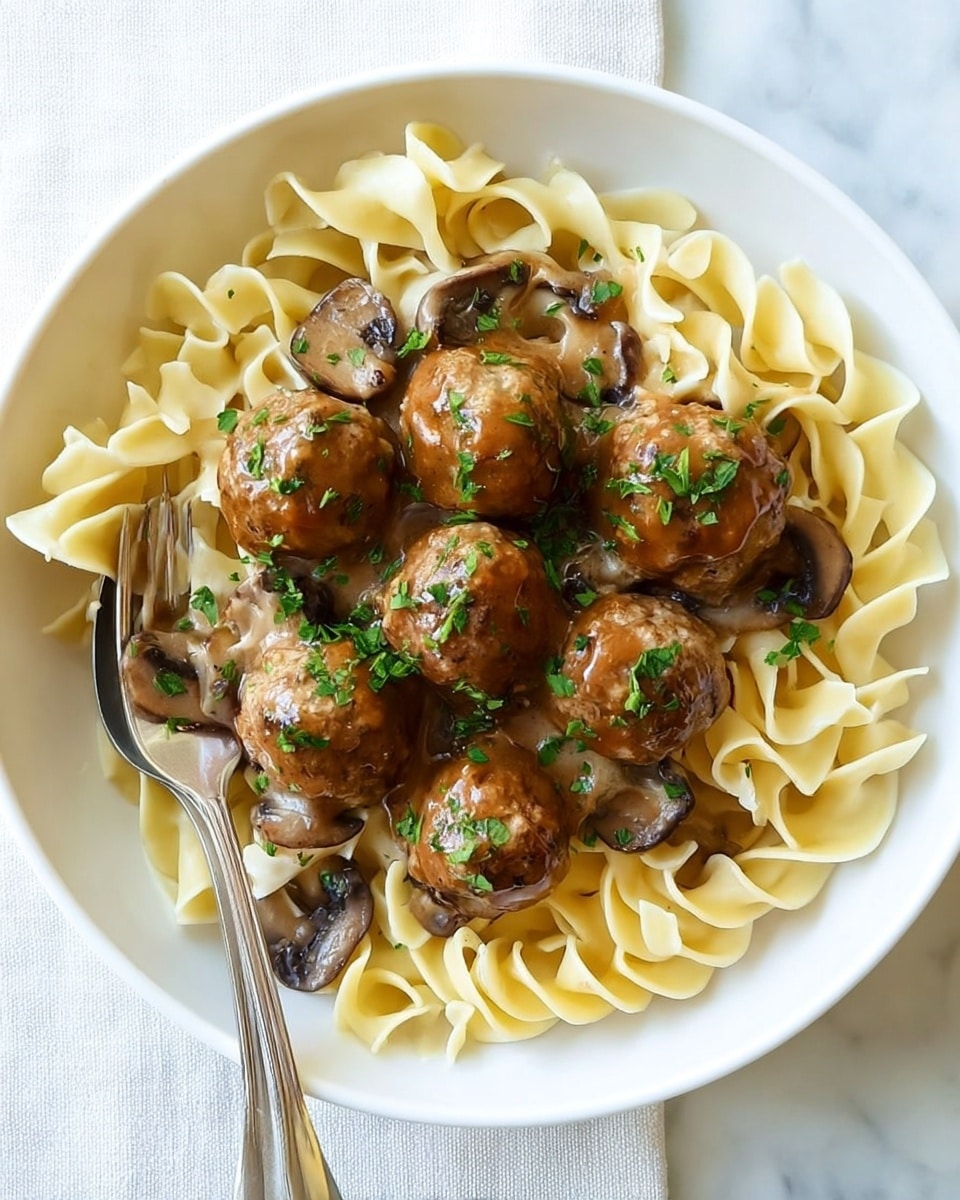 The dish shows four brown meatballs sitting on a bed of yellow buttered egg noodles with ruffled edges on a white plate with a scalloped rim. A creamy beige sauce with pale mushroom slices is drizzled over the noodles and meatballs, sprinkled with small green parsley leaves. The plate rests on a white marbled surface with a fork in the upper left corner. The lighting highlights the juicy texture of the meatballs and the smooth sauce flowing over the noodles photo taken with an iphone --ar 4:5 --v 7
