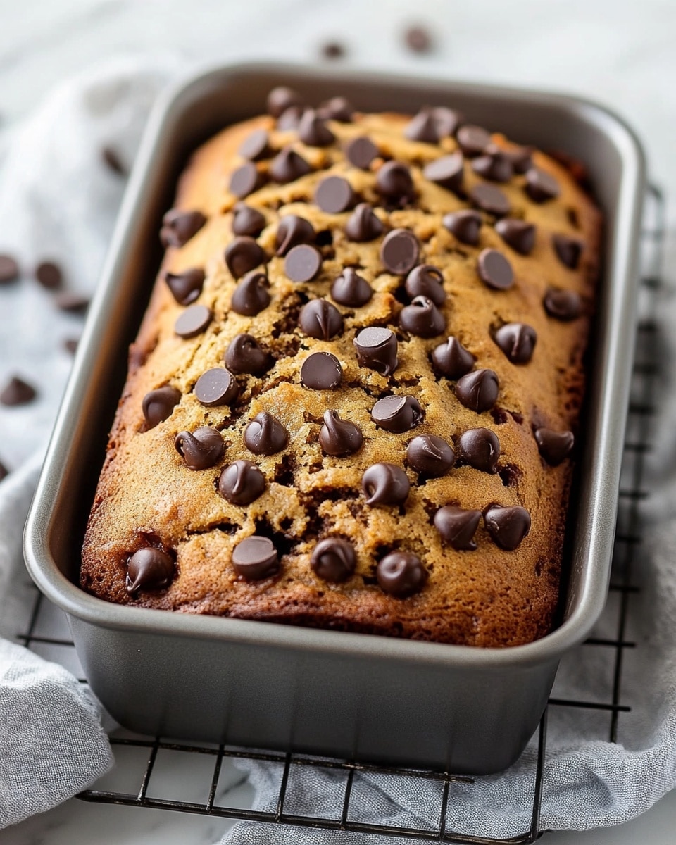 A loaf of golden brown cake baked in a grey metal rectangular pan, topped with many shiny, dark chocolate chips scattered all over the cracked, textured surface where the cake puffed up. The pan rests on a light grey cloth with a dark blue stripe, which is placed on a wire cooling rack over a white marbled surface. A few chocolate chips lie scattered blurred in the background. Photo taken with an iphone --ar 4:5 --v 6.1