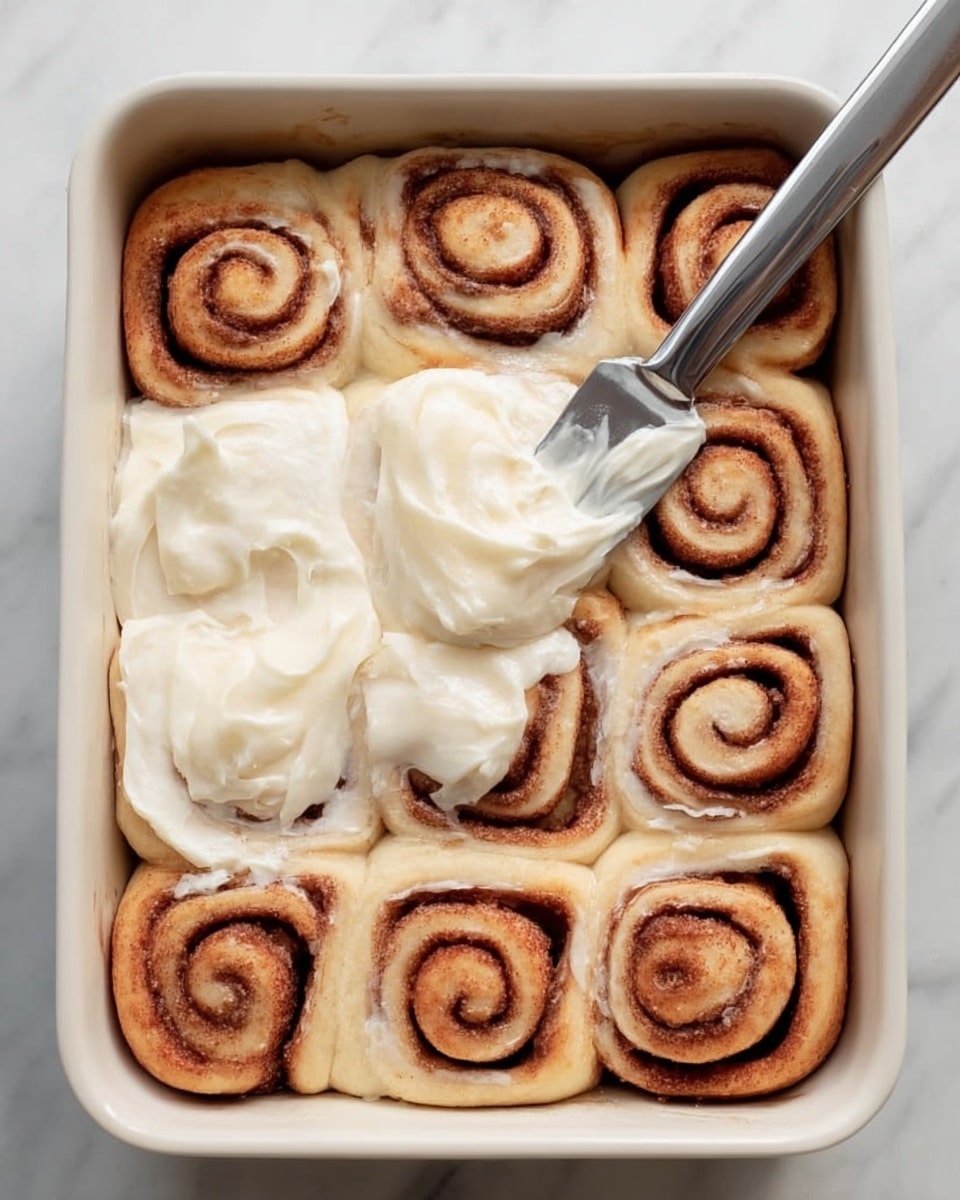 There is a white rectangular baking dish filled with 12 cinnamon rolls arranged in three rows of four. Each roll has a light golden-brown dough with darker brown cinnamon swirls in a spiral pattern. A layer of thick, creamy white frosting is being spread over the middle rolls with a knife held by a woman's hand. The dish is placed on a white marbled surface with a soft blue cloth to the side. Photo taken with an iphone --ar 4:5 --v 7