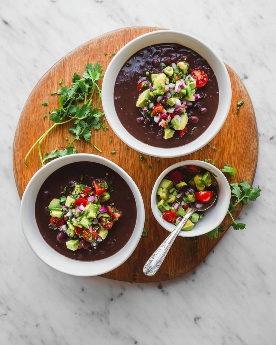 The image shows two white bowls of dark black bean soup, each topped with a colorful mixture of diced green avocado, red cherry tomatoes, chopped purple onions, and small green herbs. The bowls are placed on a round wooden board that is surrounded by fresh green cilantro sprigs. Between the two soup bowls, there is a small white bowl filled with the same avocado and tomato mixture, with a white spoon resting inside. One of the bowls has a silver spoon placed on its edge. The entire setup is on a white marbled surface. photo taken with an iphone --ar 4:5 --v 7