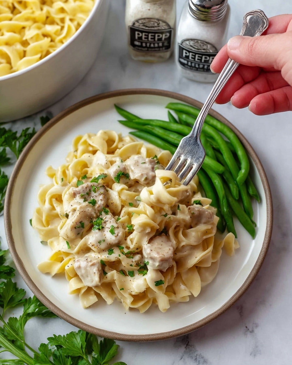 A white plate on a white marbled surface holds a serving of creamy egg noodles with pieces of light beige cooked meat mixed in, topped with small green herb flakes and light black pepper specks. To one side of the noodles, there is a small pile of bright green cooked green beans. A woman's hand holding a silver fork lifts a forkful of noodles and meat from the plate. In the background, two white shakers labeled “PEPPER” and “SALT” are slightly out of focus, along with a white bowl filled with more noodles. Fresh green parsley leaves are placed near the bottom of the plate. Photo taken with an iphone --ar 4:5 --v 7