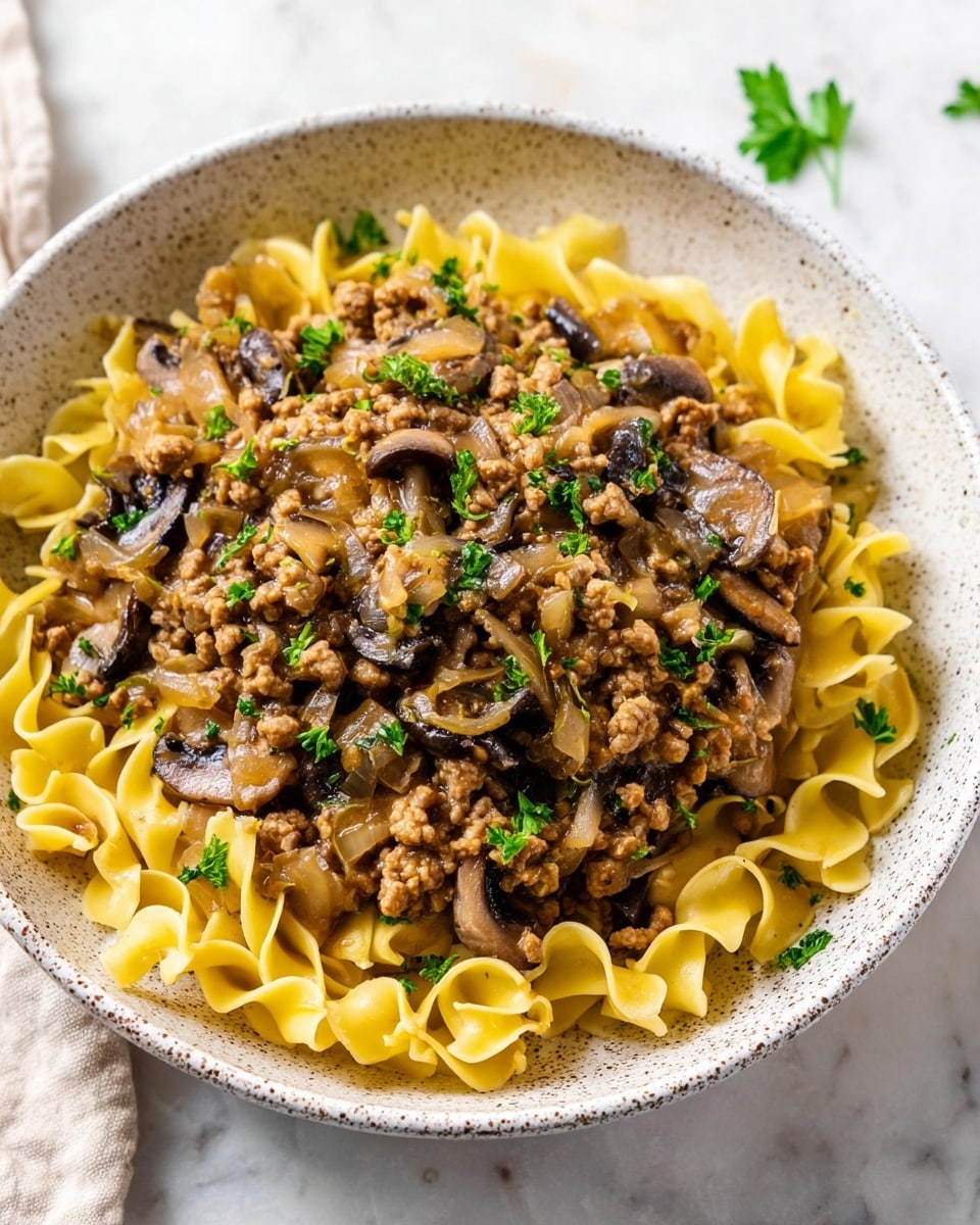 The dish is served in a white speckled bowl, placed on a white marbled surface. It has two main layers: the bottom layer is a bed of yellow egg noodles with a soft, curled texture. The top layer is a mixture of light brown, crumbly ground meat-like pieces, cooked mushrooms that are dark brown and slightly shiny, and translucent sautéed onions. This topping is mixed with a light brown sauce that moistens the ingredients. Small green parsley leaves are scattered on top for color contrast. Photo taken with an iphone --ar 4:5 --v 7
