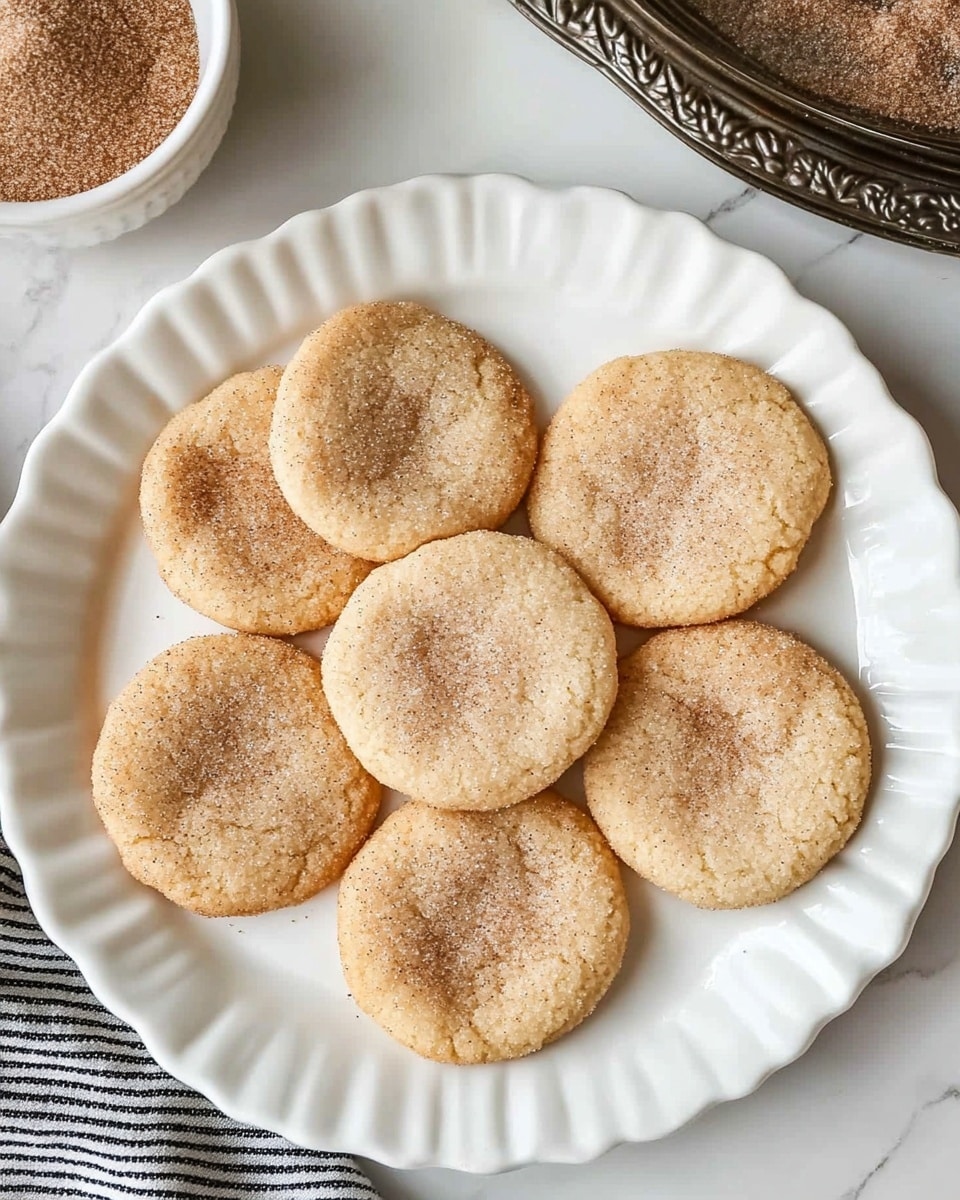 A white plate with scalloped edges holds seven round cookies arranged in a circle with one cookie in the center. The cookies are golden brown with a slightly cracked surface and are coated in a light layer of cinnamon sugar, giving a sparkling texture on top. In the upper left corner, there is a small white bowl filled with cinnamon sugar. The scene is set on a white marbled surface with a black and white striped cloth near the bottom left corner and a patterned metal tray in the upper right corner. photo taken with an iphone --ar 4:5 --v 6.1