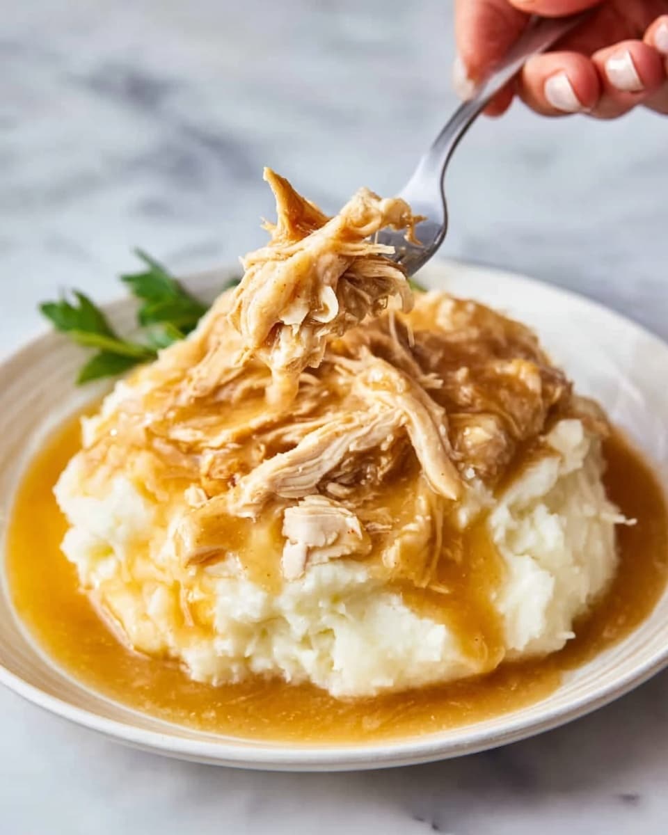 A white plate holds a thick layer of creamy white mashed potatoes with a smooth texture, topped with shredded light brown chicken covered in thick, golden brown gravy. A woman's hand is lifting some of the chicken with a silver fork, showing the soft texture of the meat and sauce. The background is a white marbled texture. photo taken with an iphone --ar 4:5 --v 7