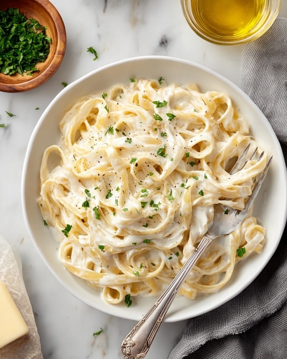 A white plate holds a serving of creamy fettuccine pasta with thick, flat noodles covered evenly in a smooth white sauce, sprinkled with small pieces of green herbs and cracked black pepper. A silver fork is placed in the pasta, resting on the plate’s right side. Above the plate to the right, there is a white pan full of the same pasta with creamy sauce and herbs. Around the plate on a white marbled surface, there is a small white bowl with pepper, a small bowl with golden olive oil, a wooden bowl with chopped green herbs, a silver cheese grater with a block of pale yellow cheese, two silver forks, and a gray cloth napkin. The scene looks bright and clean, showing different textures from creamy sauce to smooth pasta and finely chopped herbs. photo taken with an iphone --ar 4:5 --v 7