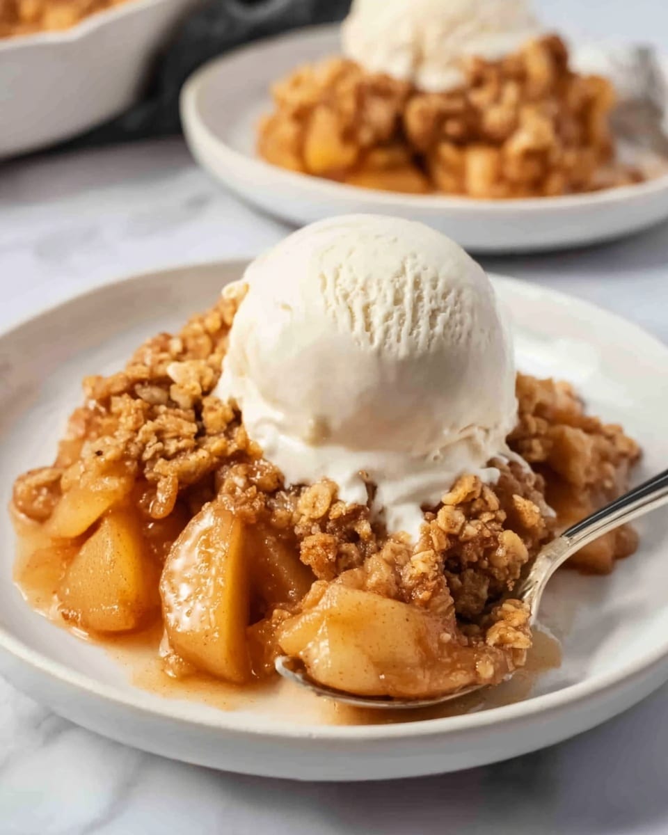 A white plate on a white marbled surface holds a warm dessert with two main layers. The bottom layer is chunky cooked apples in a shiny caramel sauce, showing soft and slightly translucent pieces. The top layer is a thick, crumbly golden-brown topping with a rough texture. On top of the crumble is a large scoop of smooth, creamy, off-white ice cream. A spoon rests on the right side of the plate, partly in the dessert, with a woman's hand holding it. In the background, a similar plate with the same dessert is partially visible, slightly out of focus. Photo taken with an iphone --ar 4:5 --v 7