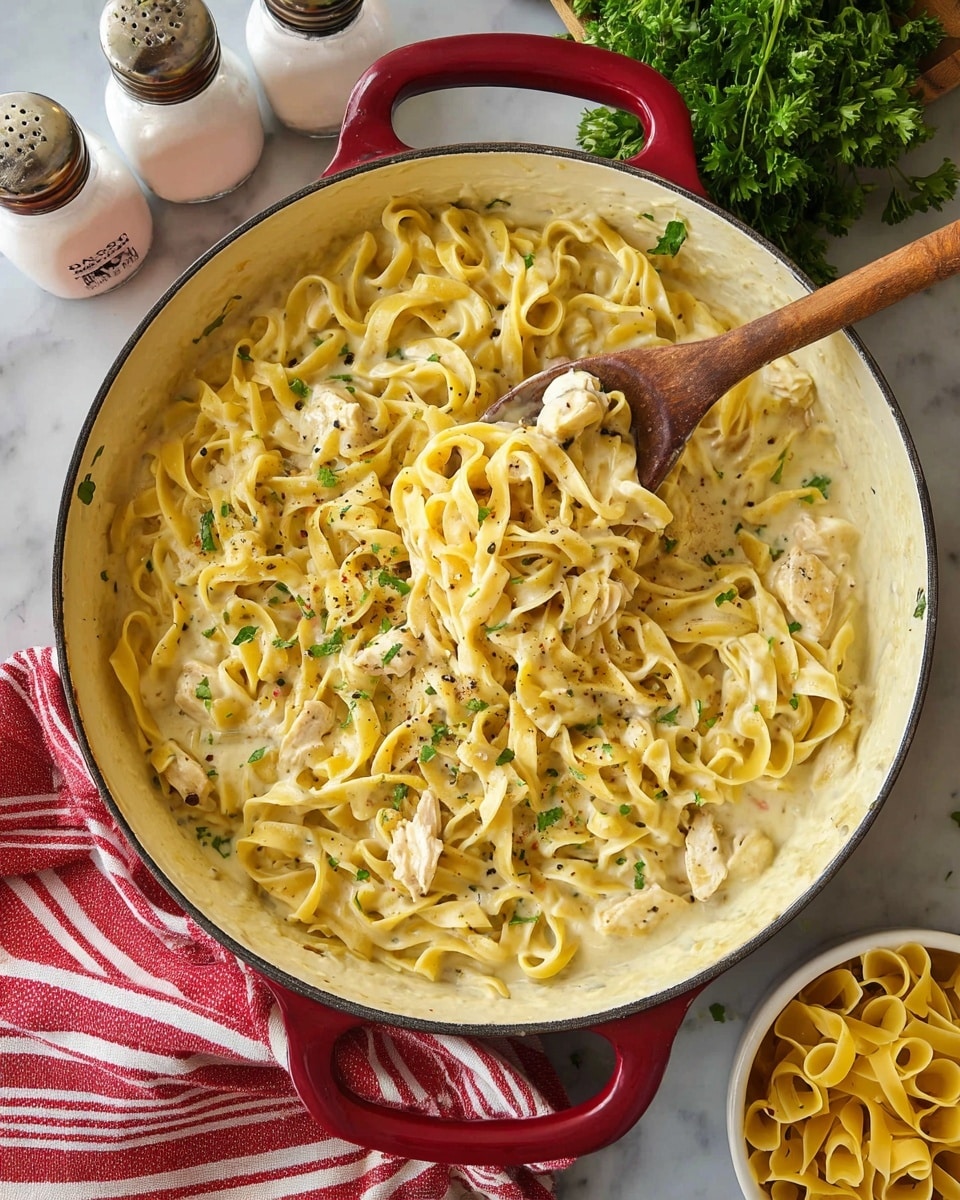 The image shows a white pan with red handles filled with creamy pasta. The pasta is yellow with a smooth, slightly shiny texture, mixed with small pieces of light-colored chicken and sprinkled with green herbs and black pepper. A wooden spoon with a rounded handle is resting inside the pan, lifting some pasta to reveal the creamy sauce beneath. Around the pan, there are two white containers labeled salt and pepper, a small bunch of fresh green parsley, a white bowl with extra uncooked pasta, and a red-striped cloth lying on a white marbled surface. photo taken with an iphone --ar 4:5 --v 7