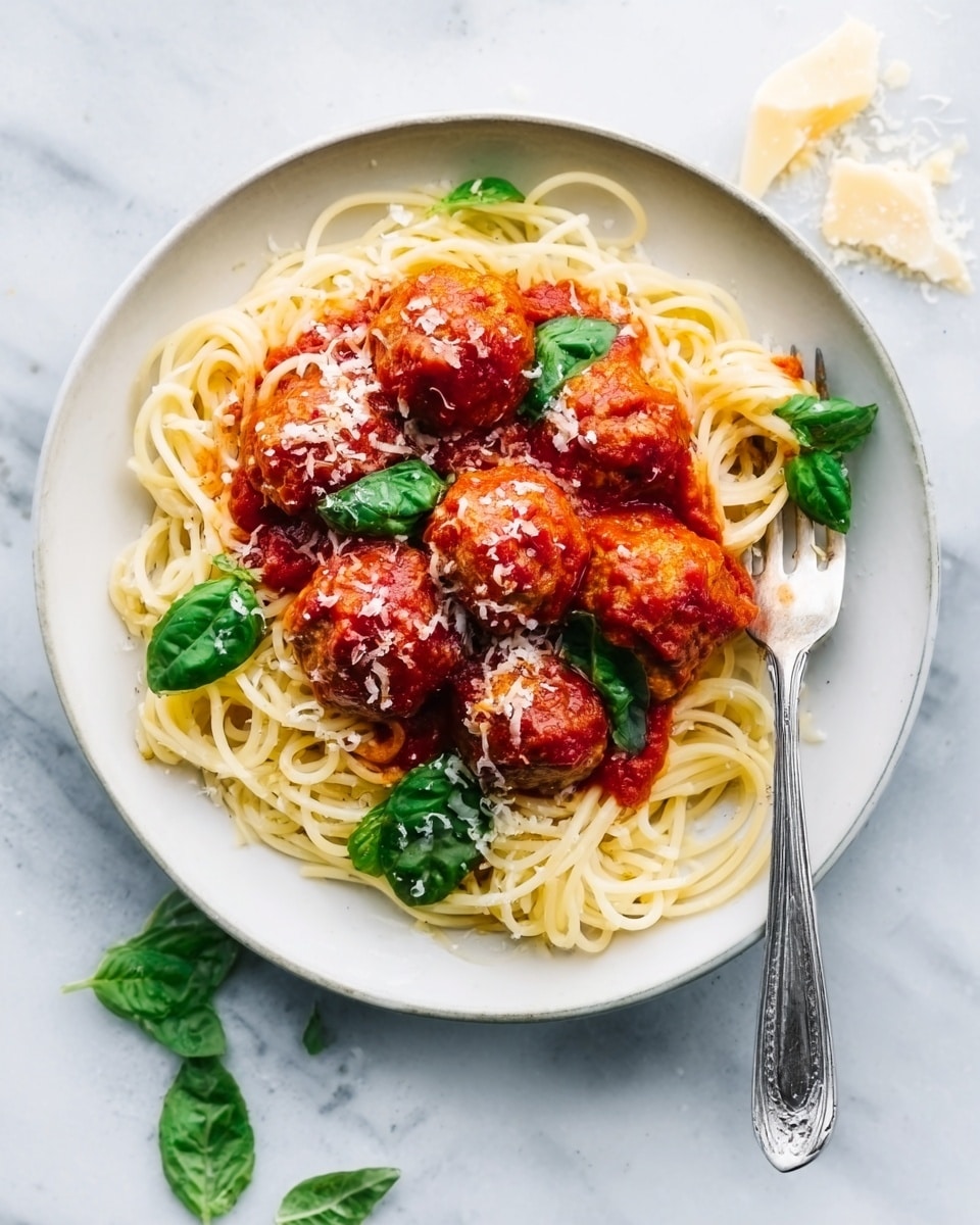 A white round plate on a white marbled surface holds a serving of spaghetti pasta arranged in a loose circular pile. On top of the pasta, there are five large red tomato-covered meatballs in a rich red tomato sauce, with a few fresh green basil leaves scattered around for color contrast. Some grated cheese is lightly sprinkled over the meatballs and sauce. A silver fork rests on the right side of the plate, with its handle extending outward. Small pieces of grated cheese and a few basil leaves are placed nearby on the surface. Photo taken with an iphone --ar 4:5 --v 7