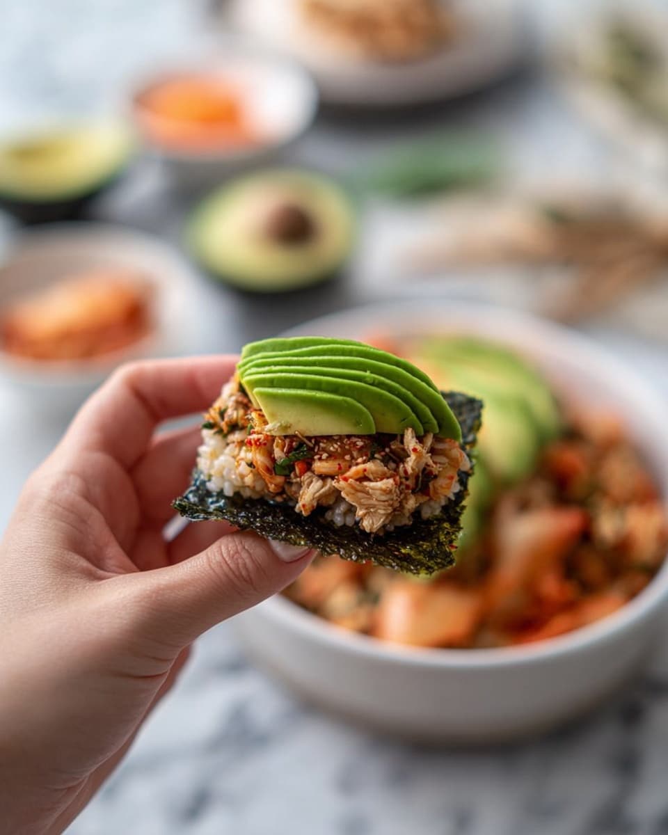 A white bowl holds two main layers: the bottom layer is a mix of lightly cooked rice with small pieces of cooked salmon, showing a mix of off-white and light pinkish orange colors. On top of this, five thin slices of vibrant green avocado are neatly fanned on one side. Small sprinkles of dark green seasoning are scattered over the rice. Surrounding the bowl on a white marbled surface are a white plate with a light pink portion of cooked salmon, a small white dish with pale yellow sesame seeds and a small silver spoon, a square white plate holding three shiny dark green seaweed sheets, and a small round dish containing reddish-orange kimchi. A gray striped cloth is partially visible near the bottom left of the scene. Photo taken with an iphone --ar 4:5 --v 7