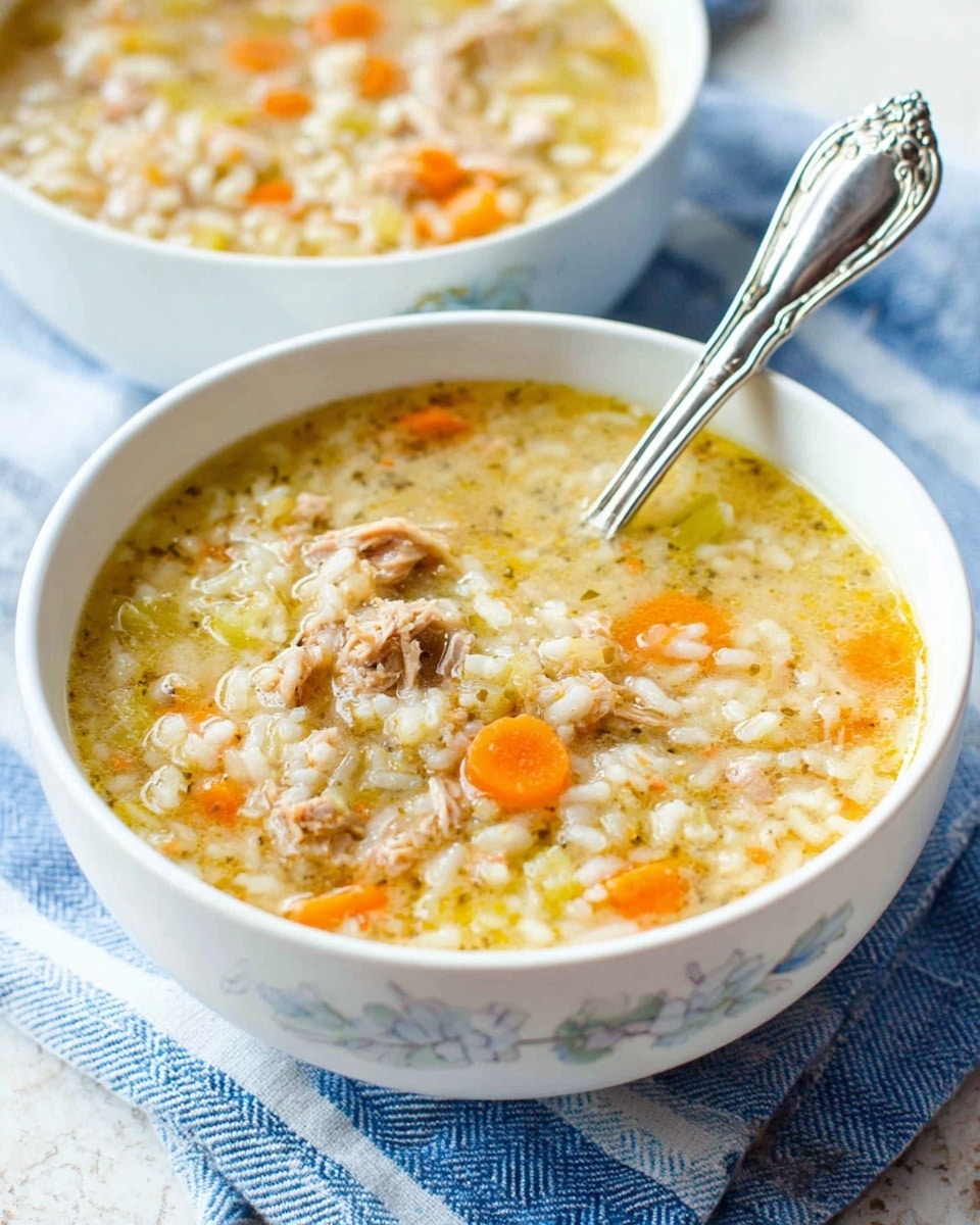 A white bowl filled with a thick soup made of shredded light brown meat, small chopped orange carrots, small white grains of rice, and light green celery bits floating in a creamy, light yellow broth. The bowl has a subtle floral design near the top edge. A silver spoon rests inside the bowl, shiny with a detailed handle. The bowl is placed on a white marbled textured surface with a blue and white striped cloth underneath, and a second bowl with the same soup is slightly blurred in the background. Photo taken with an iphone --ar 4:5 --v 7