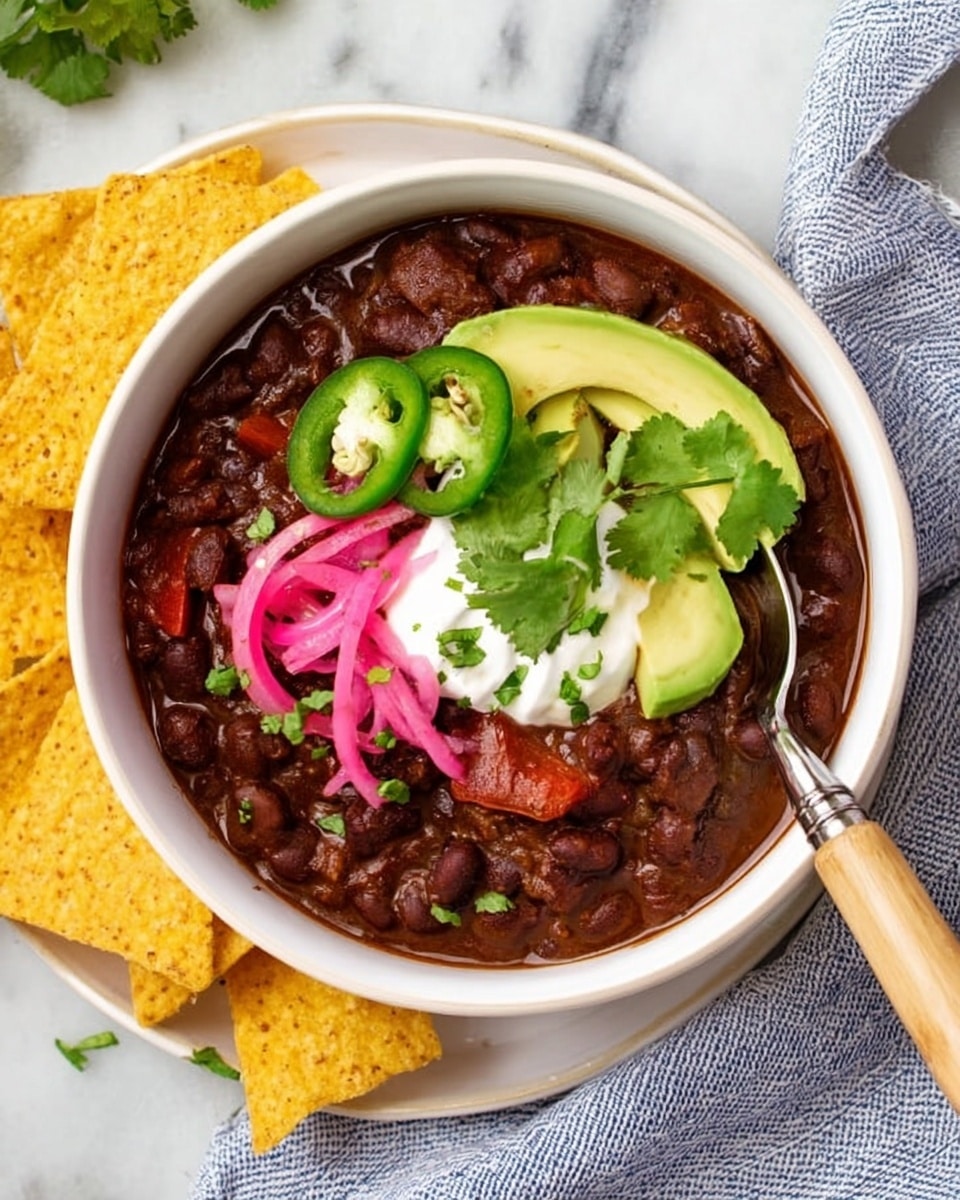 A white bowl filled with dark brown black bean chili that looks thick and chunky with pieces of red bell pepper visible. On top of the chili, there are three slices of light green avocado, a dollop of white sour cream, three thin slices of bright green jalapeño, a small bunch of fresh green cilantro leaves, and a swirl of pink pickled onions. The bowl has a silver spoon with a wooden handle resting inside. The bowl is placed on a white marbled surface with blue striped cloth nearby, and two large triangular yellow corn chips rest next to the bowl. photo taken with an iphone --ar 4:5 --v 7