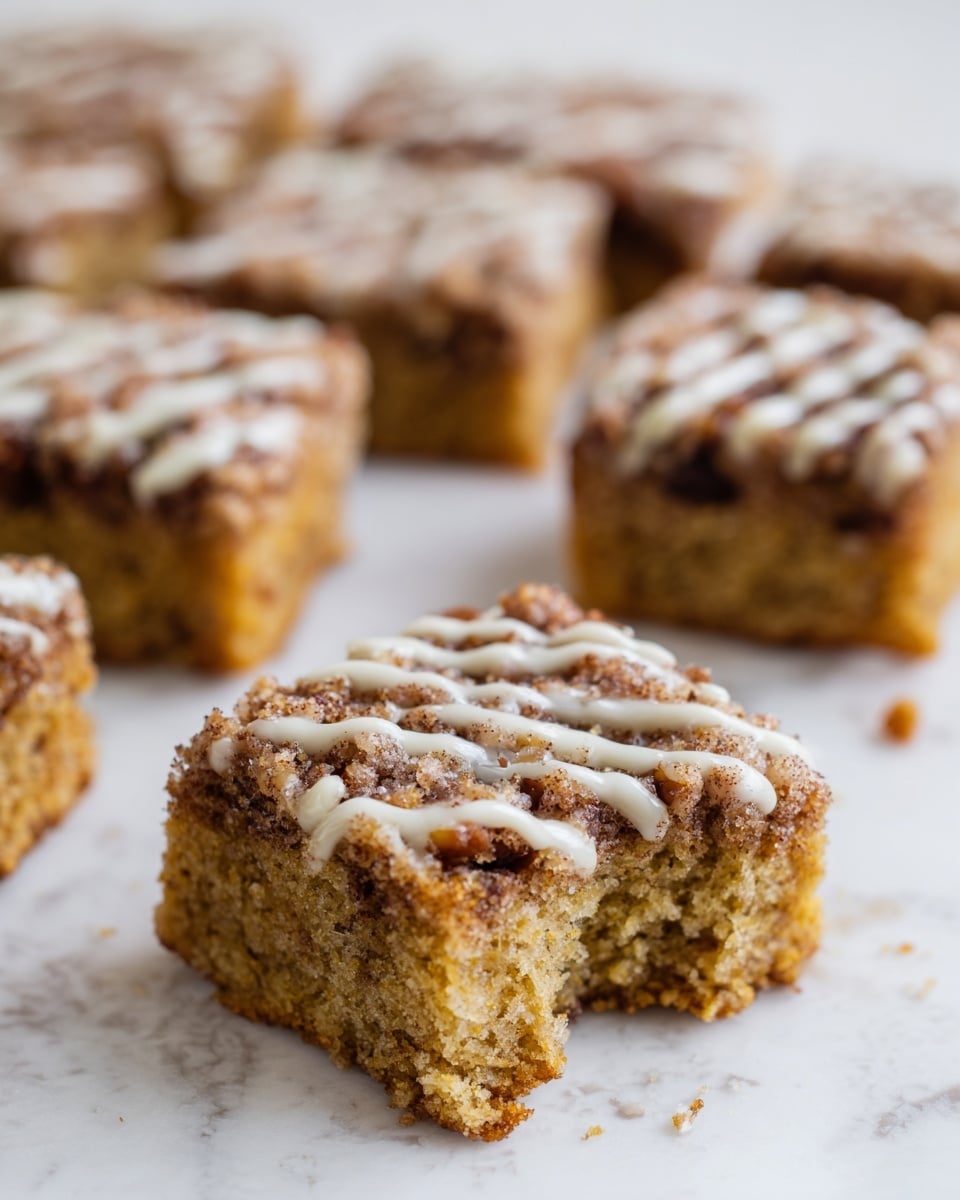 The image shows small square pieces of crumb cake with a light brown soft texture inside, and a darker brown crumbly topping mixed with small nut pieces. The top is drizzled with a thin white glaze in an uneven pattern. One piece in the front has a small bite taken out, showing its airy inside. The cake squares sit on a surface with a white marbled texture. In the background, more pieces of the crumb cake are arranged in rows. The lighting is bright and natural, showing texture clearly, photo taken with an iphone --ar 4:5 --v 7