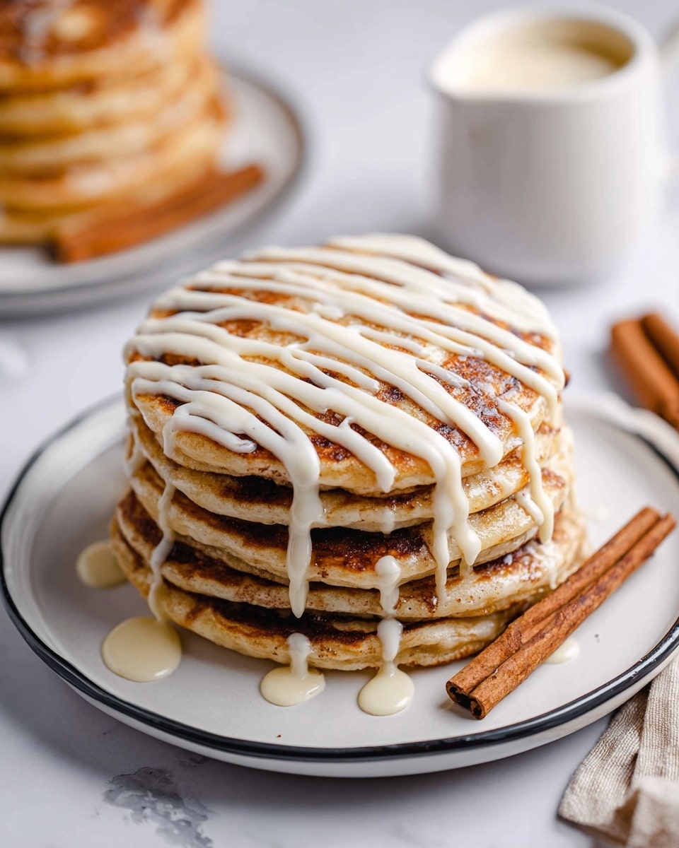 A stack of six light brown pancakes with swirled darker brown cinnamon layers visible between each one sits on a white plate with a thin black rim, placed on a white marbled surface. The top pancake is decorated with white icing drizzled in even horizontal lines across the stack. A slice has been lifted by a silver fork showing the fluffy texture and the cinnamon swirl inside. In the background, another stack of pancakes is partially visible with similar white icing drizzled on top, next to a small white jug filled with cream or sauce. photo taken with an iphone --ar 4:5 --v 7