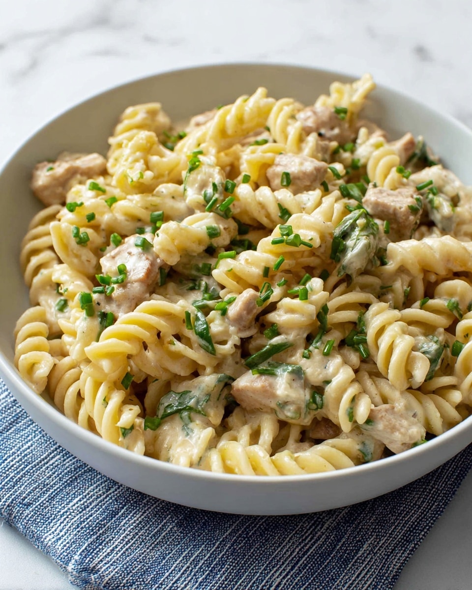 A white bowl filled with creamy rotini pasta mixed with pieces of light beige cooked chicken, bright green spinach leaves, and small bits of onion. The creamy sauce coats the pasta and chicken evenly giving a smooth light yellowish color. Chopped green herbs are sprinkled on top for a fresh touch. The bowl is set on a white marbled surface next to a gray cloth with white stripes. photo taken with an iphone --ar 4:5 --v 7