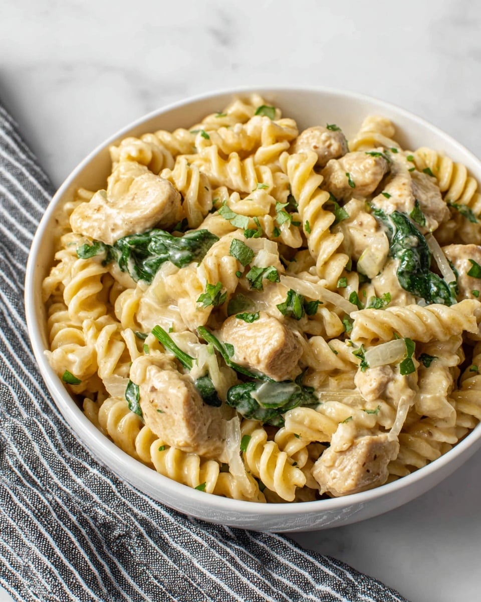 A white bowl filled with creamy pasta made of spiral rotini in a pale yellow sauce. Mixed in are chunks of light brown cooked chicken and vibrant green spinach leaves, scattered evenly throughout the dish. Small pieces of green herbs are sprinkled on top for added color. The bowl sits on a white marbled surface beside a gray and white striped cloth. Photo taken with an iphone --ar 4:5 --v 7