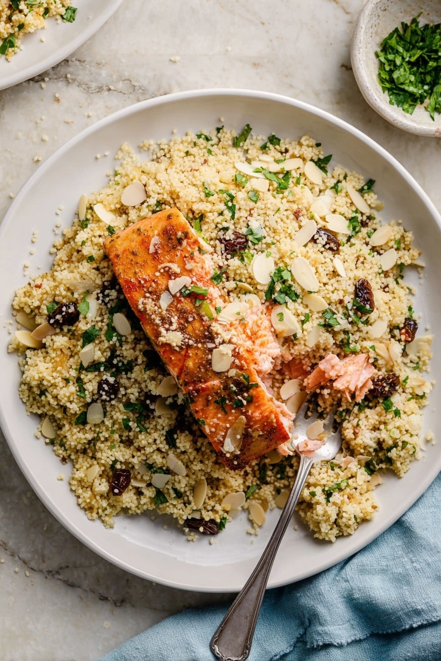 A white bowl filled with a base layer of light yellow couscous mixed with chopped green herbs and small dark raisins. On top of the couscous, there is a piece of cooked salmon with a golden brown crust, broken open to show the soft pink inside. Thin almond slices and extra chopped green herbs are scattered on the dish. A silver spoon rests on the right side of the bowl, partially under the couscous. The bowl sits on a white marbled surface, and a light blue cloth napkin is visible nearby. photo taken with an iphone --ar 2:3 --v 7 - 30-Minute Moroccan-Spiced Salmon and Couscous, Moroccan-Spiced Salmon, quick salmon recipes, healthy seafood dinner, easy Moroccan-inspired meals