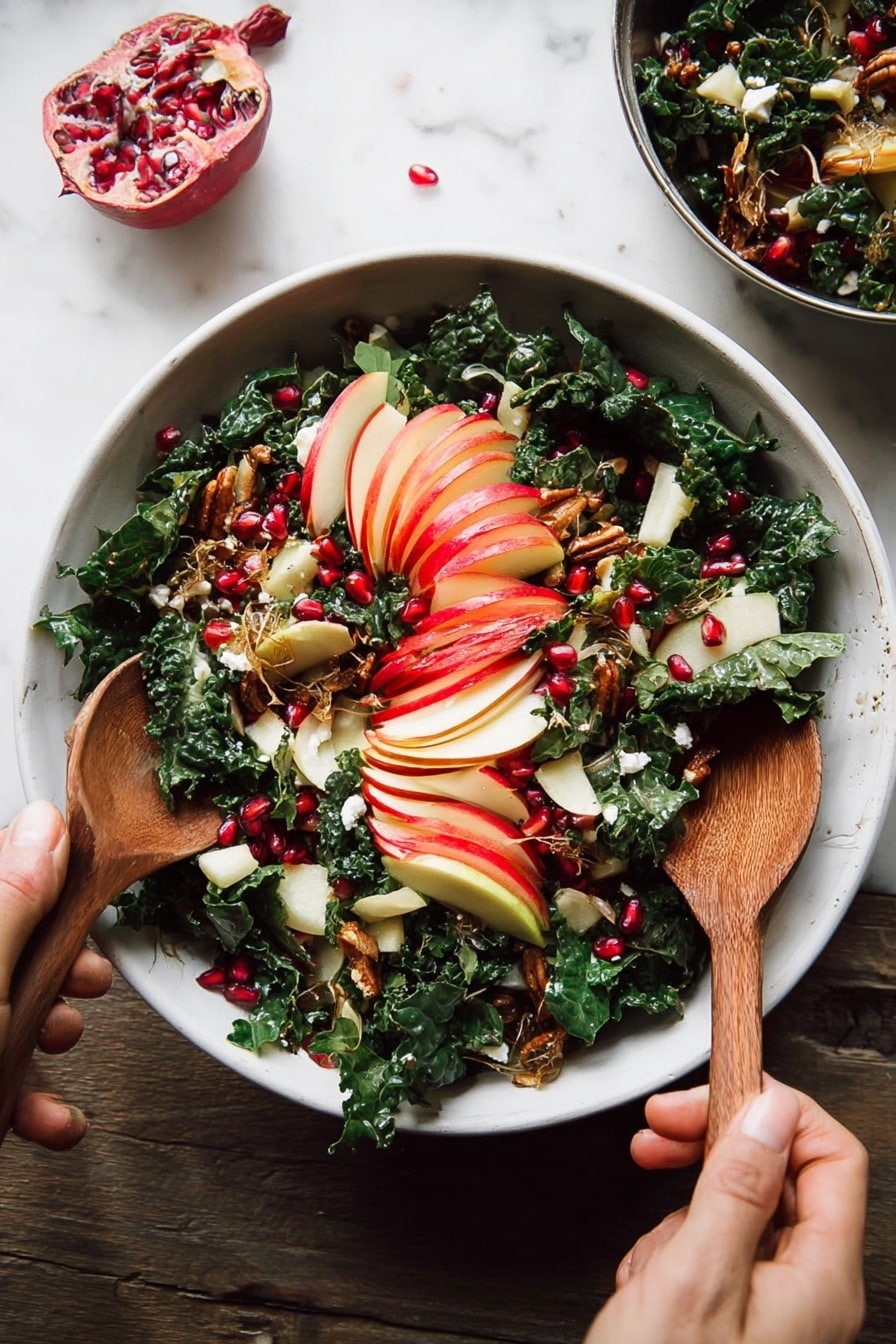 A white bowl filled with a fresh salad rests on a white marbled surface, showing layers of dark green chopped kale mixed with thin slices of red-skinned apple arranged in a fan shape on top. Bright red pomegranate seeds are scattered across the salad, adding spots of vibrant color. Golden brown toasted seeds and small bits of crispy brown topping are sprinkled on the salad for texture. Pieces of white cheese with a soft texture are visible beneath the kale. A woman's hand holds a wooden spoon on the bottom left while another woman's hand grips a second wooden spoon on the top right as they mix the salad. Photo taken with an iphone --ar 2:3 --v 7 - Fall Harvest Honeycrisp Apple and Kale Salad, autumn apple kale salad, healthy fall salad, seasonal apple and kale salad, caramelized shallot vinaigrette salad