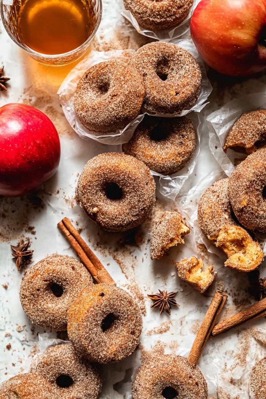 The image shows many cinnamon sugar donuts placed closely on a white marbled surface. Most donuts are whole, with a rough texture covered in sugar and cinnamon. A few donuts are broken, revealing a soft, golden inside with a slightly crumbly texture. Some donuts are wrapped in thin, translucent white paper, adding layers and depth. Near the donuts, there is a bright red apple and a clear glass cup filled with light brown tea and cinnamon sticks inside. Star anise is scattered around for decoration. The scene looks warm and cozy with soft natural light. Photo taken with an iphone --ar 2:3 --v 7 - Baked Cinnamon Crunch Apple Cider Doughnuts, fall baked doughnuts, cinnamon sugar doughnuts, apple cider baked treats, easy autumn doughnut recipe