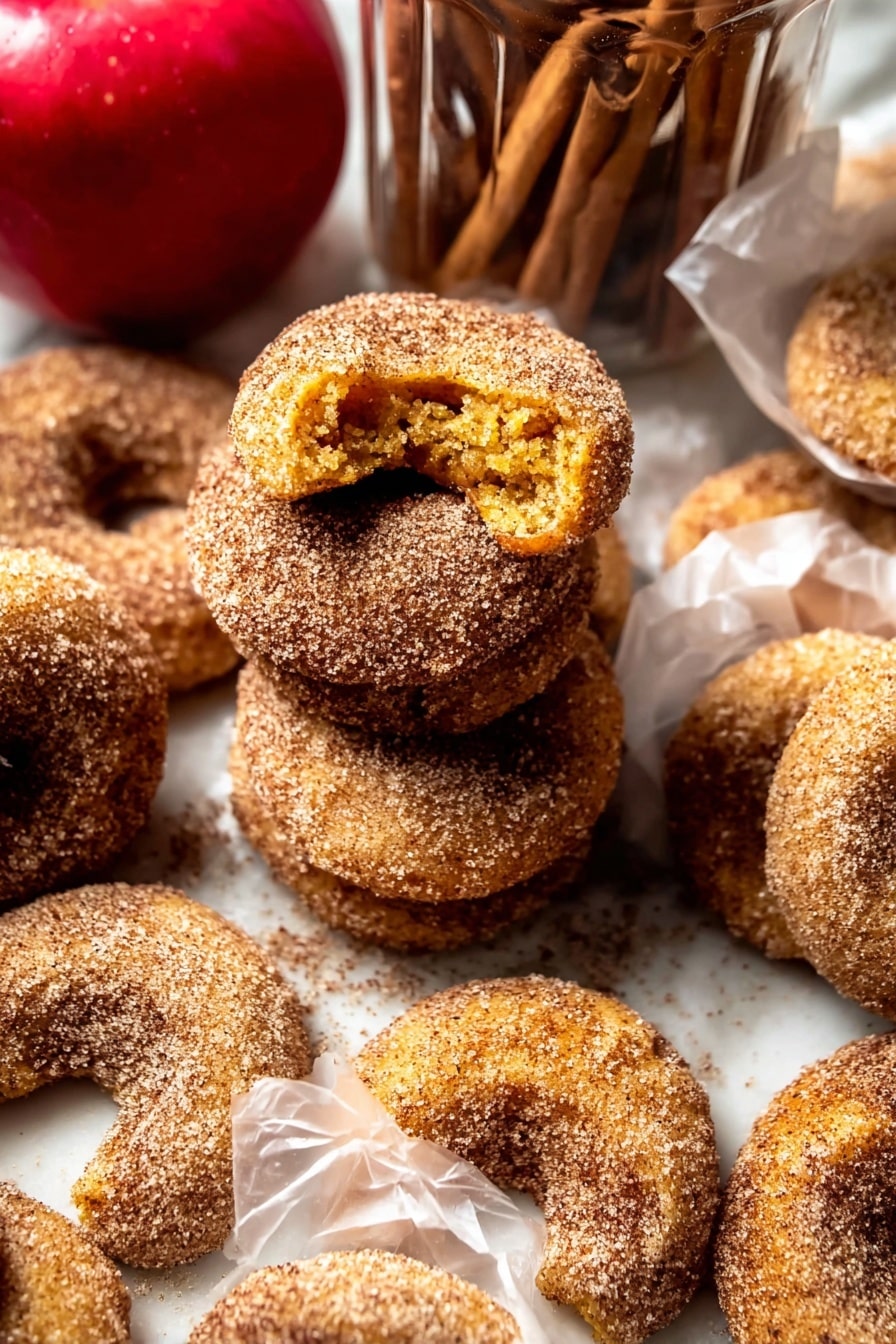 The image shows several crescent-shaped cookies covered in a coarse layer of cinnamon sugar. The cookies are golden brown with a slightly rough texture beneath the sugar coating. Some cookies are stacked in a loose pile, while others are individually wrapped in translucent white paper. The cookies have a crumbly look, with one cookie broken open to reveal a soft yellow interior. In the background, there is a bright red apple and a clear glass jar filled with more cookies and cinnamon sticks on top. The setting uses a white marbled surface underneath all the items. photo taken with an iphone --ar 2:3 --v 7 - Baked Cinnamon Crunch Apple Cider Doughnuts, fall baked doughnuts, cinnamon sugar doughnuts, apple cider baked treats, easy autumn doughnut recipe