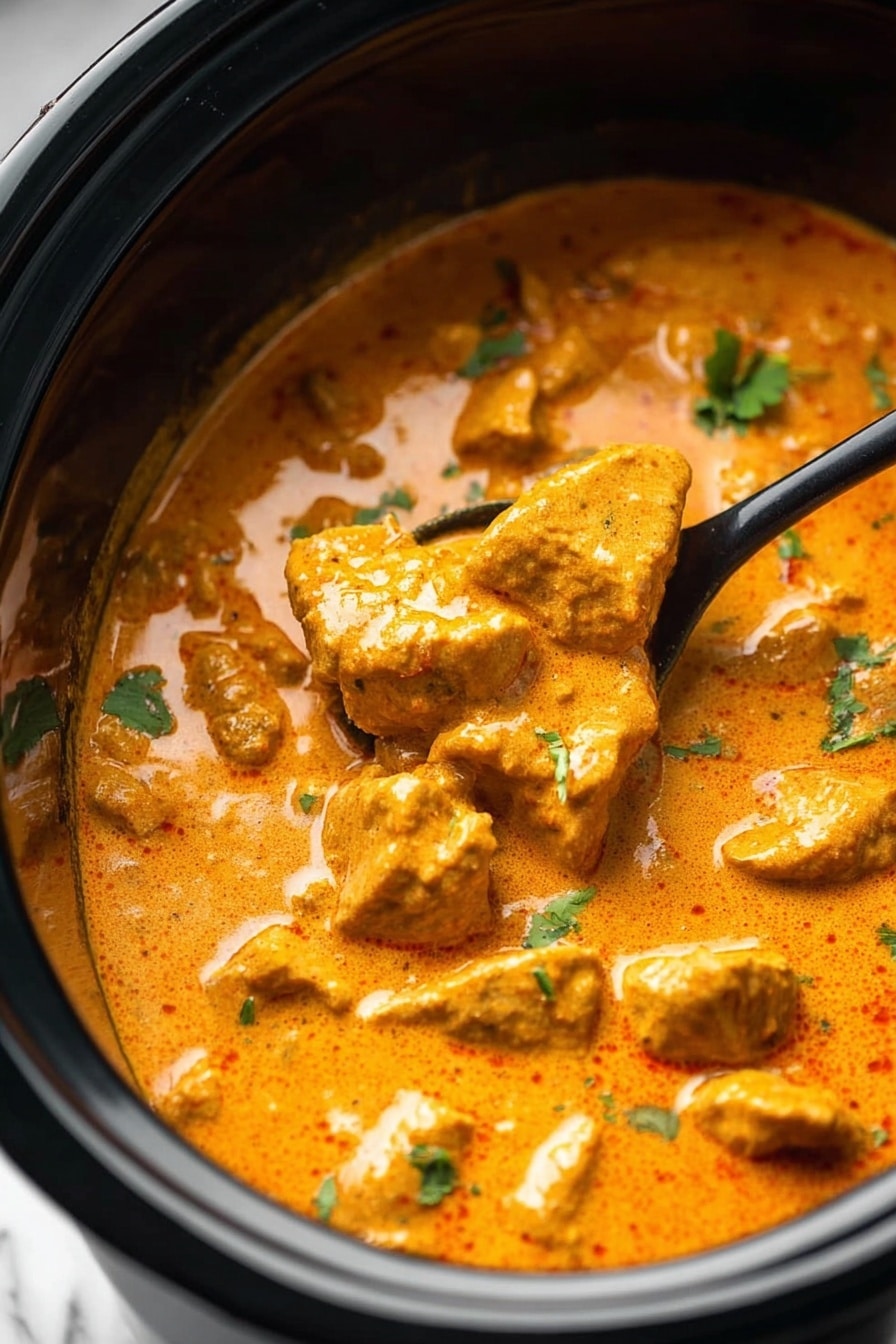 This image shows a close-up of a creamy orange curry with chunks of tender meat in a black slow cooker. The curry sauce is thick and smooth with a slightly shiny surface. Small green herb pieces are sprinkled on top, adding a fresh contrast to the warm orange sauce. A black spoon is partially submerged in the curry on the right side, lifting some pieces of the meat. The background is a white marbled texture. photo taken with an iphone --ar 2:3 --v 7 - Slow Cooker Butter Chicken, Easy Butter Chicken, Creamy Chicken Curry, Make-Ahead Chicken Dinner, Instant Pot Butter Chicken