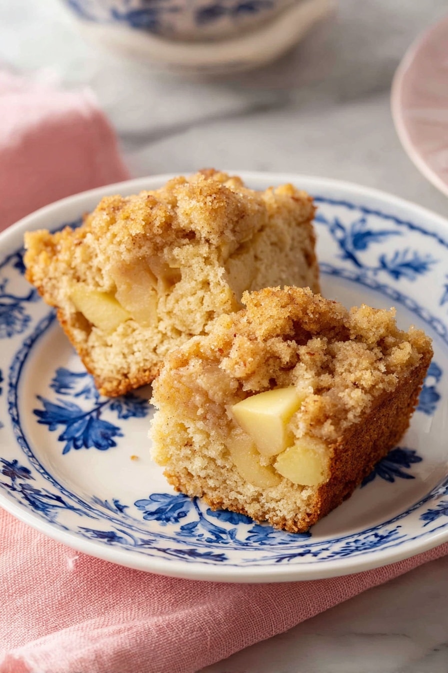 Two pieces of crumb-topped cake are placed on a white plate with blue floral patterns. The cake has a light brown color with soft, crumbly texture and visible chunks of yellow apple inside. The crumb topping is thicker and golden brown. The plate sits on a pink cloth, all set on a white marbled surface. Photo taken with an iphone --ar 2:3 --v 7 - Apple Muffins, apple muffin recipe, homemade apple muffins, cinnamon apple muffins, easy muffin recipes