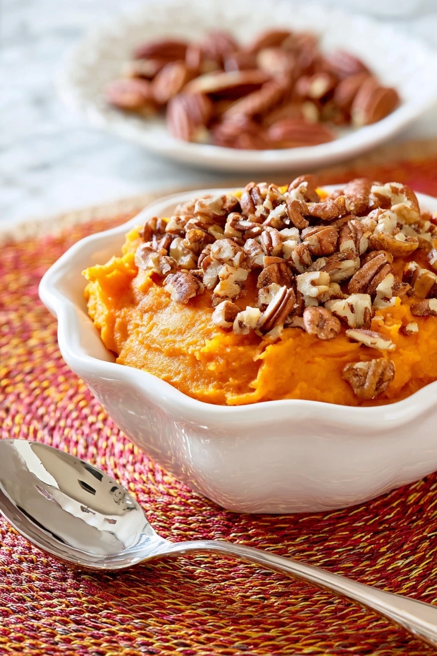 This image shows a white bowl filled with a thick, mashed orange sweet potato layer that is roughly shaped with a soft texture. On top of this layer, there is a generous amount of chopped pecans scattered unevenly, adding a crunchy contrast with their brown, nutty pieces. The bowl sits on a woven orange, red, and yellow circular placemat, and in the blurred background, part of another white dish with light brown pecans can be seen resting on a white marbled surface. photo taken with an iphone --ar 2:3 --v 7 - Slow Cooker Sweet Potato Casserole with Maple and Toasted Pecans, sweet potato casserole, slow cooker sweet potato dish, holiday sweet potato recipe, easy Thanksgiving side