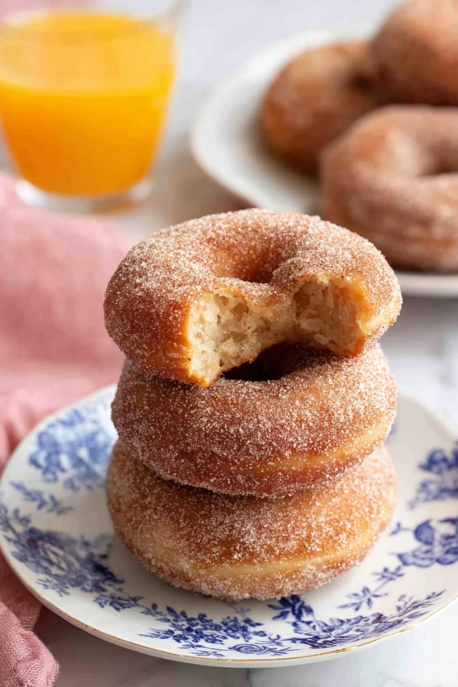 Three cinnamon sugar donuts are stacked on a white plate with blue floral designs, showing a soft texture covered in fine sugar crystals; the top donut has a bite taken, revealing a light, fluffy inside with a slightly darker core. Behind the plate is a blurred white plate with more cinnamon sugar donuts and a clear glass filled with bright orange juice, all set on a white marbled surface. A pink cloth is placed softly on the left side. photo taken with an iphone --ar 2:3 --v 7 - Super Moist Apple Cider Donuts with Warm Spices, apple cider donuts recipe, fall donuts with warm spices, easy apple cider donut recipe, cozy autumn dessert