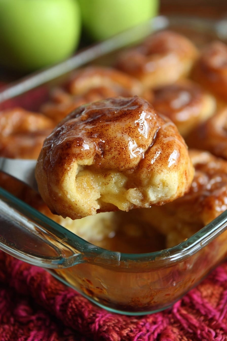 This image shows a close-up of a golden brown pastry with a shiny glaze, held above a glass baking dish filled with similar pastries. The pastry has a baked, slightly rough surface with visible cinnamon specks and a soft, filling visible at one side. The glass dish is resting on a thick, red knitted cloth, with blurred green apples in the background. photo taken with an iphone --ar 2:3 --v 7 - Apple Dumplings with Cinnamon Sugar and Sprite Drizzle, apple dumplings recipe, cinnamon sugar dessert, easy apple dessert, quick fruit dessert