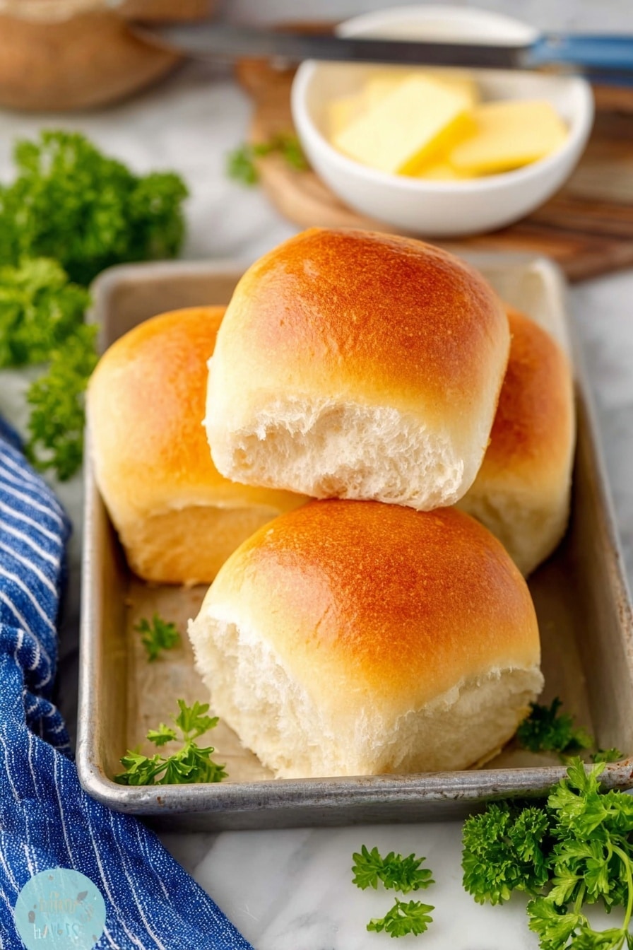 The image shows four soft, shiny bread rolls with a golden brown top, stacked and placed inside a metal baking tray on a white marbled surface. The bread looks fluffy and light, with a slightly textured white inside. Behind the rolls, there is a small white bowl holding a pale yellow stick of butter and a butter knife resting on the bowl's edge. Bright green parsley leaves are scattered around the tray, adding a fresh touch. A blue and white striped cloth is partly visible on the left side, contributing to a cozy, homey feel. photo taken with an iphone --ar 2:3 --v 7 - Homemade Fluffy Dinner Rolls, fluffy dinner rolls recipe, soft buttery dinner rolls, easy homemade dinner rolls, bakery-style dinner rolls