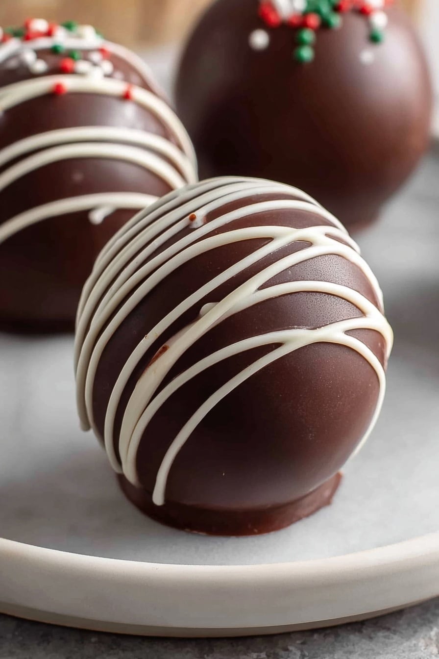 The image shows a smooth, shiny, dark brown chocolate sphere placed on a white plate with a white marbled texture under it. The sphere has thin white chocolate lines drizzled across its surface in a loose crisscross pattern, creating contrast and delicate texture on the curved chocolate shell. In the background, two more similar chocolate spheres are partially visible, each decorated with the same white drizzle and small dots of colored sprinkles. The overall look is clean and glossy, with the sphere’s curved layers clearly defined. photo taken with an iphone --ar 2:3 --v 7 - Hot Chocolate Bombs, Hot Chocolate Bombs Recipe, Easy Hot Chocolate Bombs, Homemade Hot Chocolate Bombs, Christmas Hot Chocolate Bombs