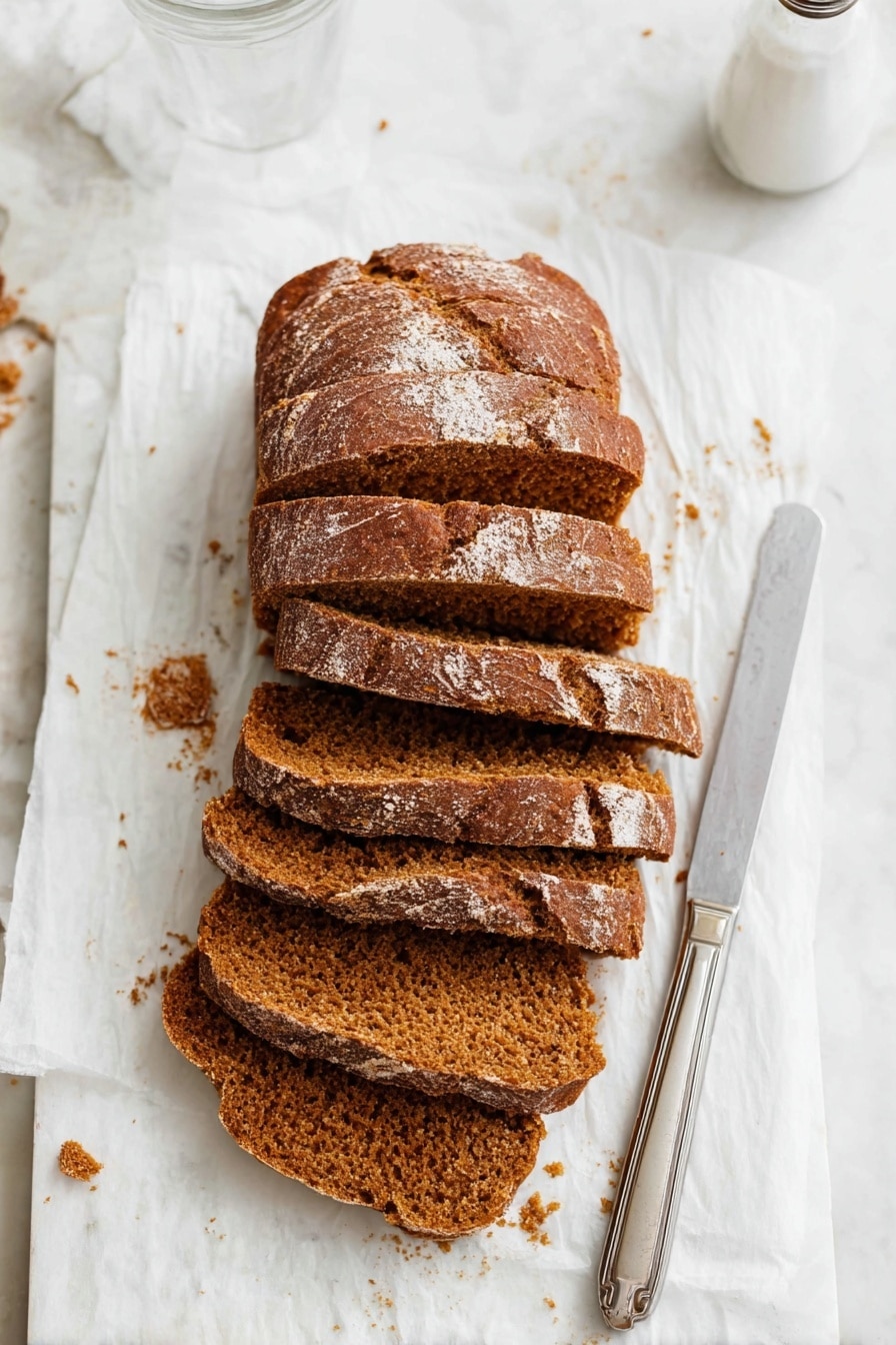 The image shows a loaf of brown bread cut into six thick slices, placed slightly overlapping each other on a white parchment paper. The bread has a textured crust with a few light flour spots on top and a soft, crumbly inside with a warm brown color. To the right of the bread, there is a silver knife with a simple handle resting on the parchment paper. The background is a white marbled surface with some bread crumbs scattered around, and the top right part of a clear glass and a white salt shaker are visible. Photo taken with an iphone --ar 2:3 --v 7 - Pumpkin Bread, Pumpkin Bread for Fall, Easy Pumpkin Bread, Moist Pumpkin Bread, Fall Pumpkin Baking