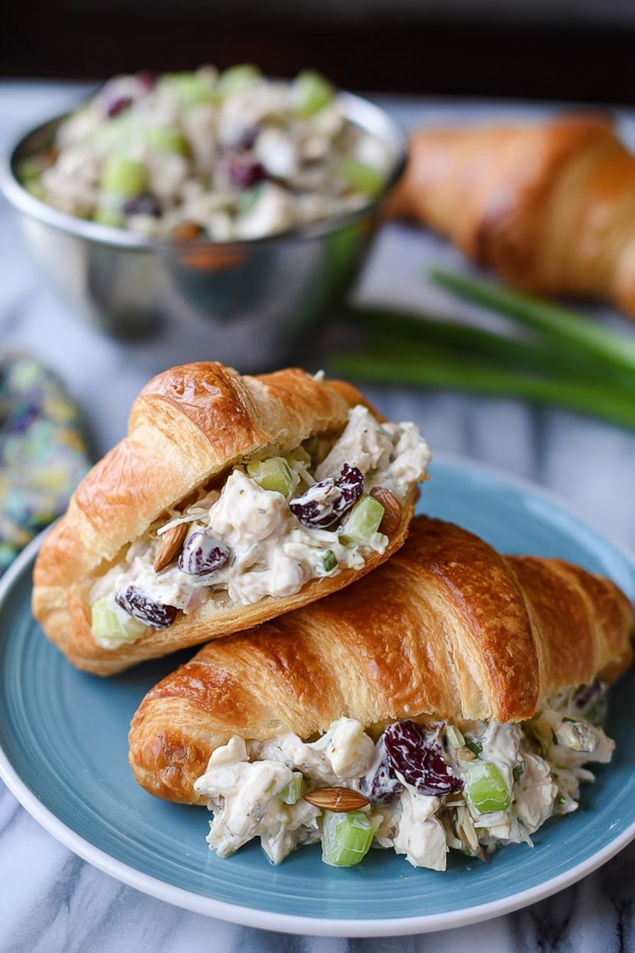 The image shows two croissants on a blue plate placed on a white marbled surface. One croissant is whole with a golden-brown, flaky texture. The other croissant is open and filled with a creamy salad made of small white chicken pieces, chopped celery, dried dark cranberries, sunflower seeds, and light beige dressing, layered inside the croissant’s light and flaky interior. In the background, there is a metal bowl filled with more of the salad, and some green onions and an additional croissant are blurred. Photo taken with an iphone --ar 2:3 --v 7 - Cranberry Chicken Salad, Cranberry Chicken Salad Recipe, Easy Chicken Salad with Cranberries, Healthy Cranberry Chicken Salad, Refreshing Chicken Salad