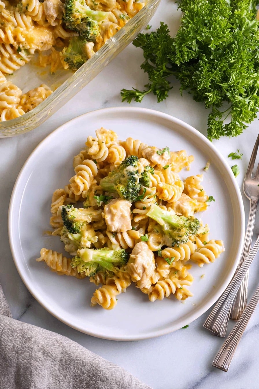 A white plate holds a serving of pasta made with spiral-shaped noodles mixed with chunks of light-colored cooked chicken and small, bright green broccoli florets, all coated in a creamy light yellow cheese sauce with a slightly shiny texture; the dish is placed on a white marbled surface next to a partially visible glass baking dish filled with more pasta, a small bunch of fresh parsley with deep green leaves, and a folded light gray cloth napkin holding three metal forks. Photo taken with an iphone --ar 2:3 --v 7 - Chicken Pasta Casserole with Broccoli, easy chicken pasta bake, cheesy chicken casserole, healthy broccoli pasta dish, family-friendly comfort food