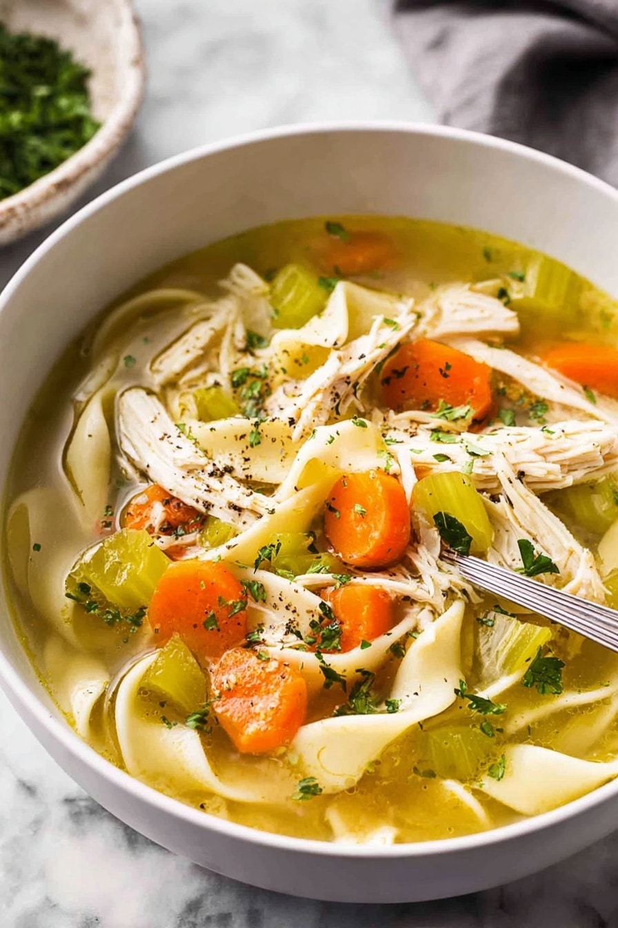 The image shows a close-up view of a white bowl filled with clear chicken soup. Inside the soup, there are wide flat noodles curled softly, bright orange carrot slices, pieces of pale green celery, and shredded white chicken meat scattered on one side. Small green parsley flakes float on the surface along with some black pepper specks. The soup is light golden and clear, with visible layers of ingredients resting naturally in the broth. The bowl sits on a white marbled surface. photo taken with an iphone --ar 2:3 --v 7 - Homemade Chicken Noodle Soup from Scratch, chicken noodle soup from scratch, homemade chicken soup, hearty chicken noodle recipe, easy homemade chicken soup