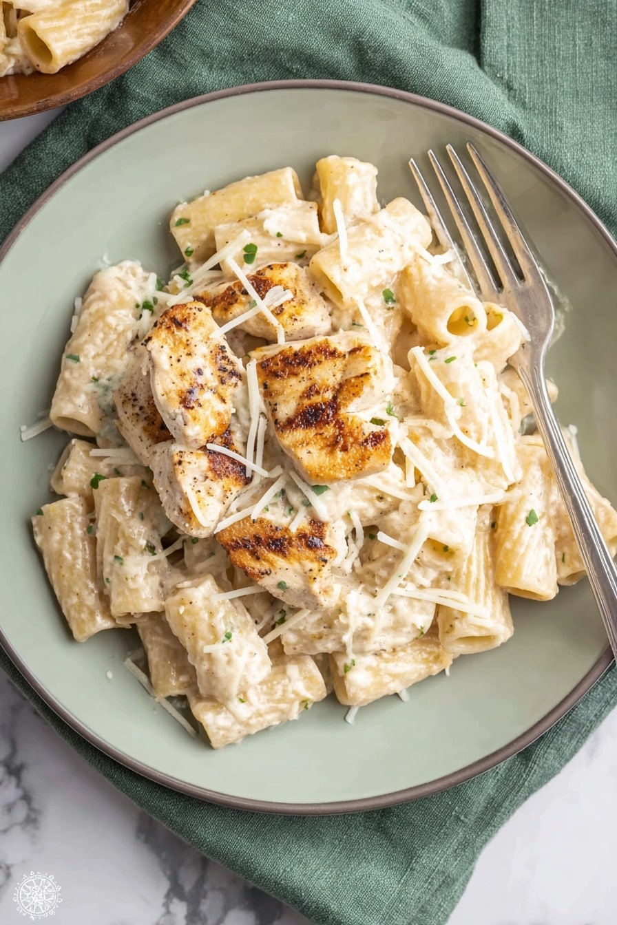 A white plate holds a serving of rigatoni pasta coated in a creamy white sauce with small green herb flecks. On top of the pasta lie four grilled chicken pieces, light brown with darker grill marks, partially covered by the sauce. Thin white shredded cheese is sprinkled over the chicken and pasta, adding texture. A silver fork rests on the right side of the plate. The plate is on a soft green cloth placed on a white marbled surface. Photo taken with an iphone --ar 2:3 --v 7 - One Pot Creamy Parmesan Chicken Pasta, creamy chicken pasta, one pot chicken pasta dinner, easy creamy pasta recipe, quick chicken pasta meal