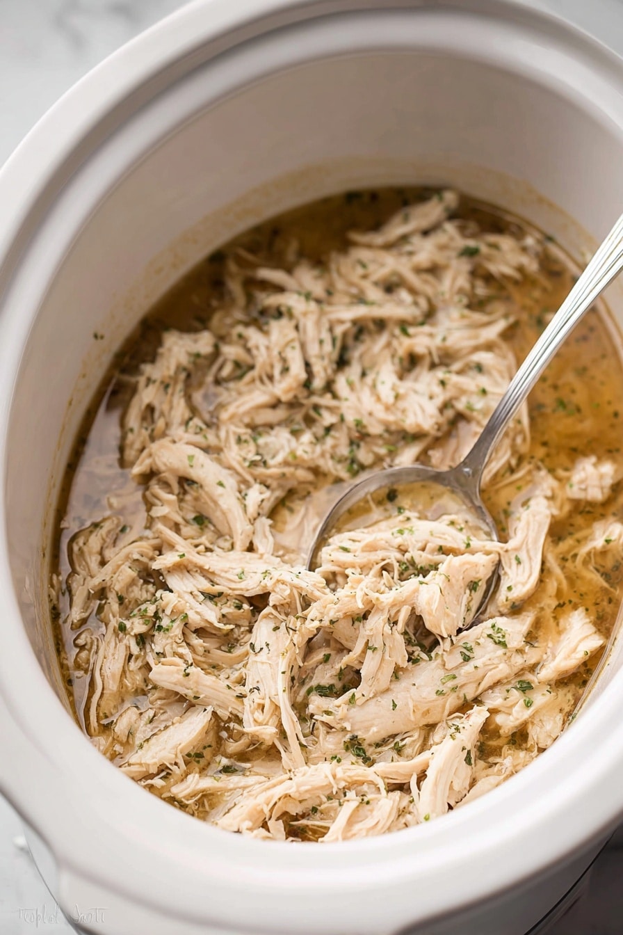 Inside a white slow cooker, there is one main layer of shredded light beige cooked chicken mixed with small green herb pieces, all soaked in a thin, light brown broth. A silver spoon rests partially submerged in the mixture on the right side. The chicken strands have a soft texture, and the herbs are finely sprinkled. The slow cooker sits on a white marbled surface. photo taken with an iphone --ar 2:3 --v 7 - Juicy Crockpot Shredded Chicken, Crockpot shredded chicken, easy shredded chicken recipes, perfect shredded chicken, tender shredded chicken